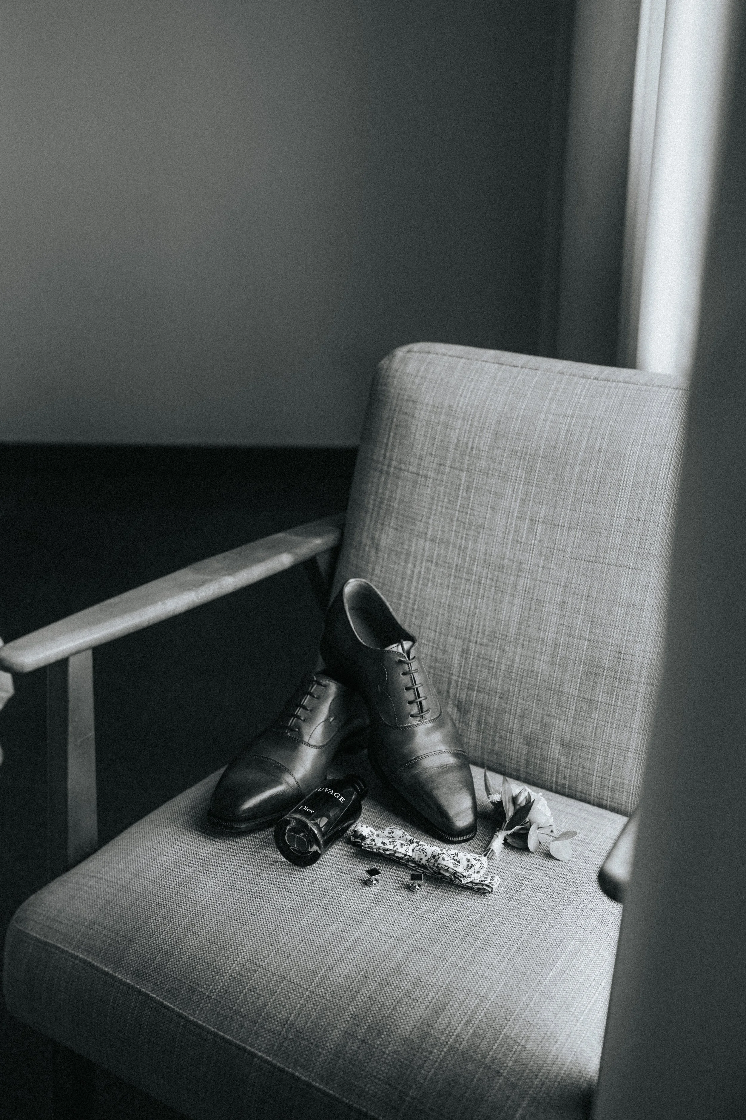 Gray upholstered chair with men's black leather dress shoes, a black coffee mug, a wrapped gift with black and white pattern, and scattered small items on the seat. A light-colored table and a part of a dark wall are visible.
