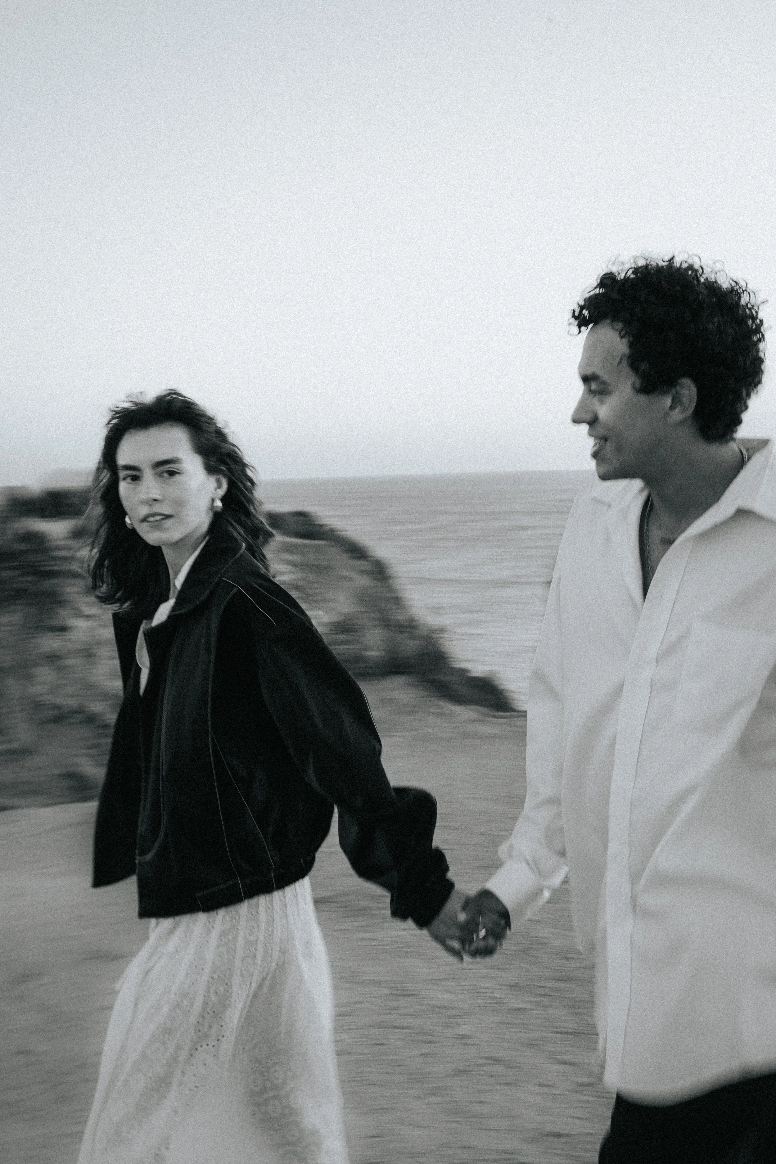 A black-and-white photo of a man and a woman holding hands while walking outdoors. The woman has dark hair, wearing earrings, a dark jacket, and a white skirt. The man has curly hair, wearing a light-colored shirt and dark pants. They are walking nea