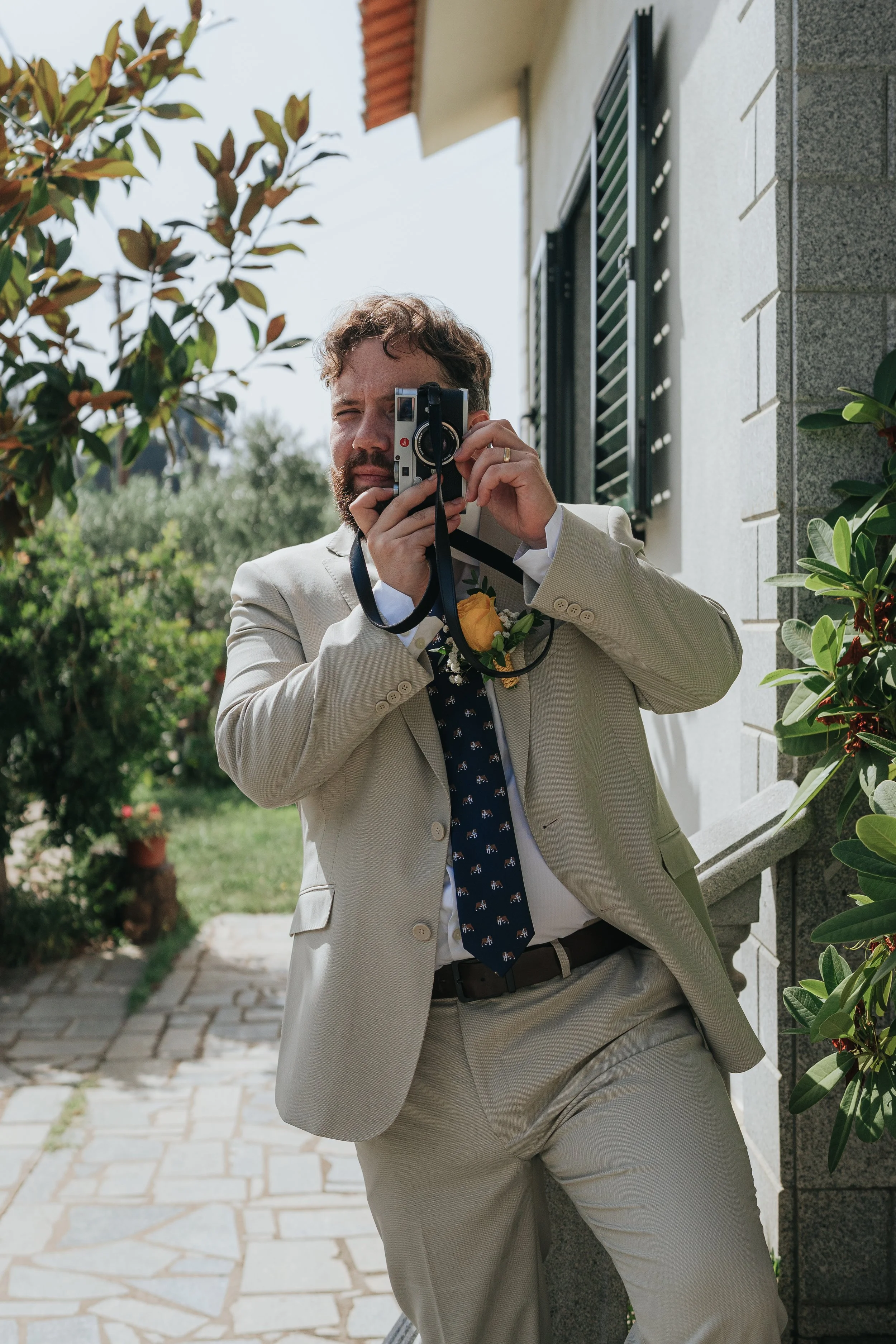 A man in a light-colored suit and dark tie taking a photo outside next to a building with greenery, holding a camera to his face.