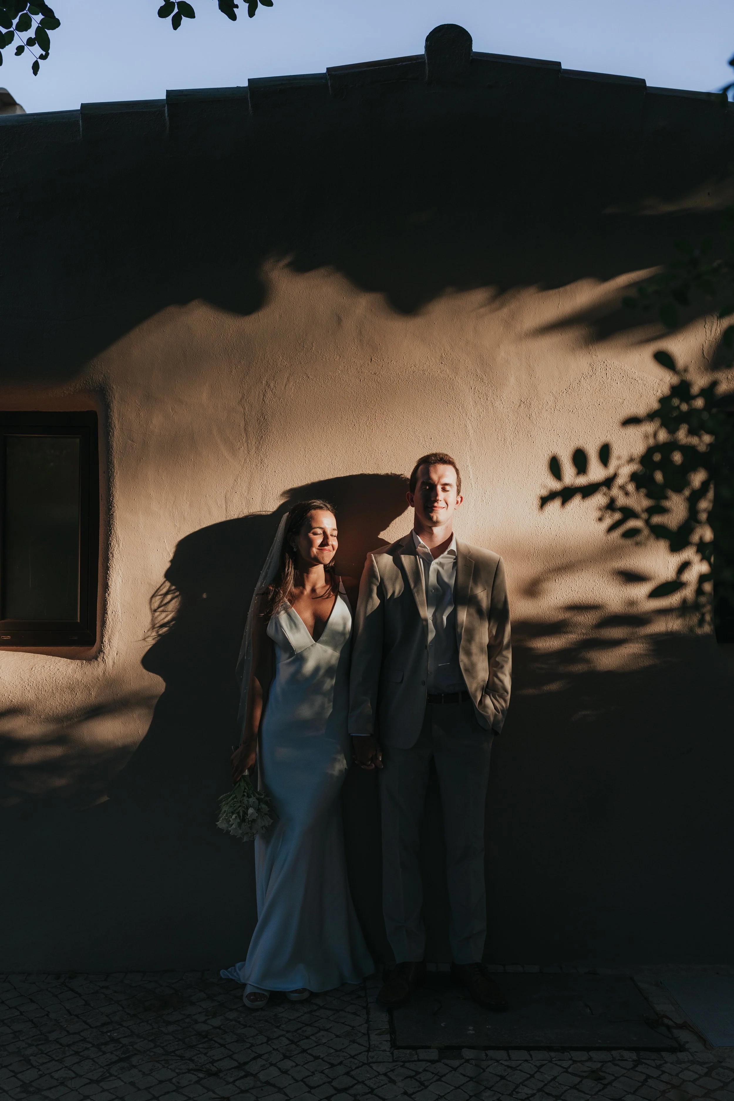A bride and groom standing outdoors against a beige stucco wall, holding hands, with shadow play and leaves framing the top and right side of the image. The bride is smiling with eyes closed, wearing a white wedding dress, and the groom is looking ah