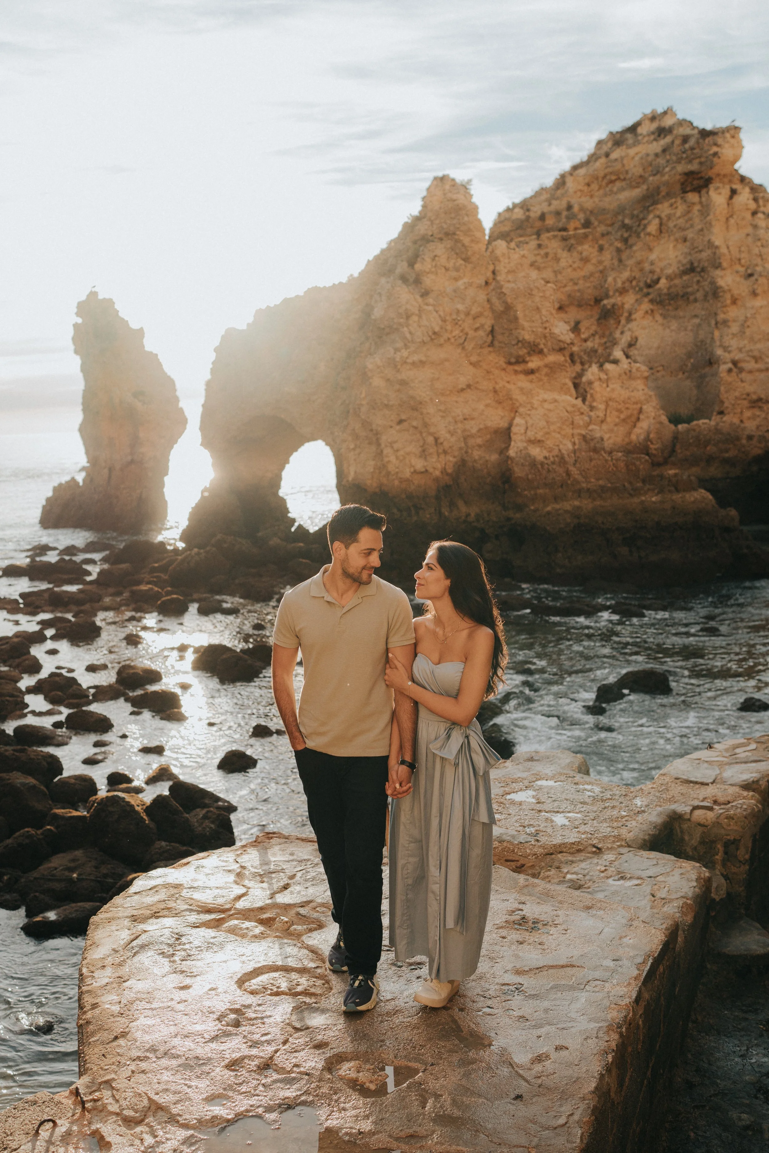 A couple holding hands and walking on a rocky ledge near the ocean, with large rock formations and arches in the background during sunset.