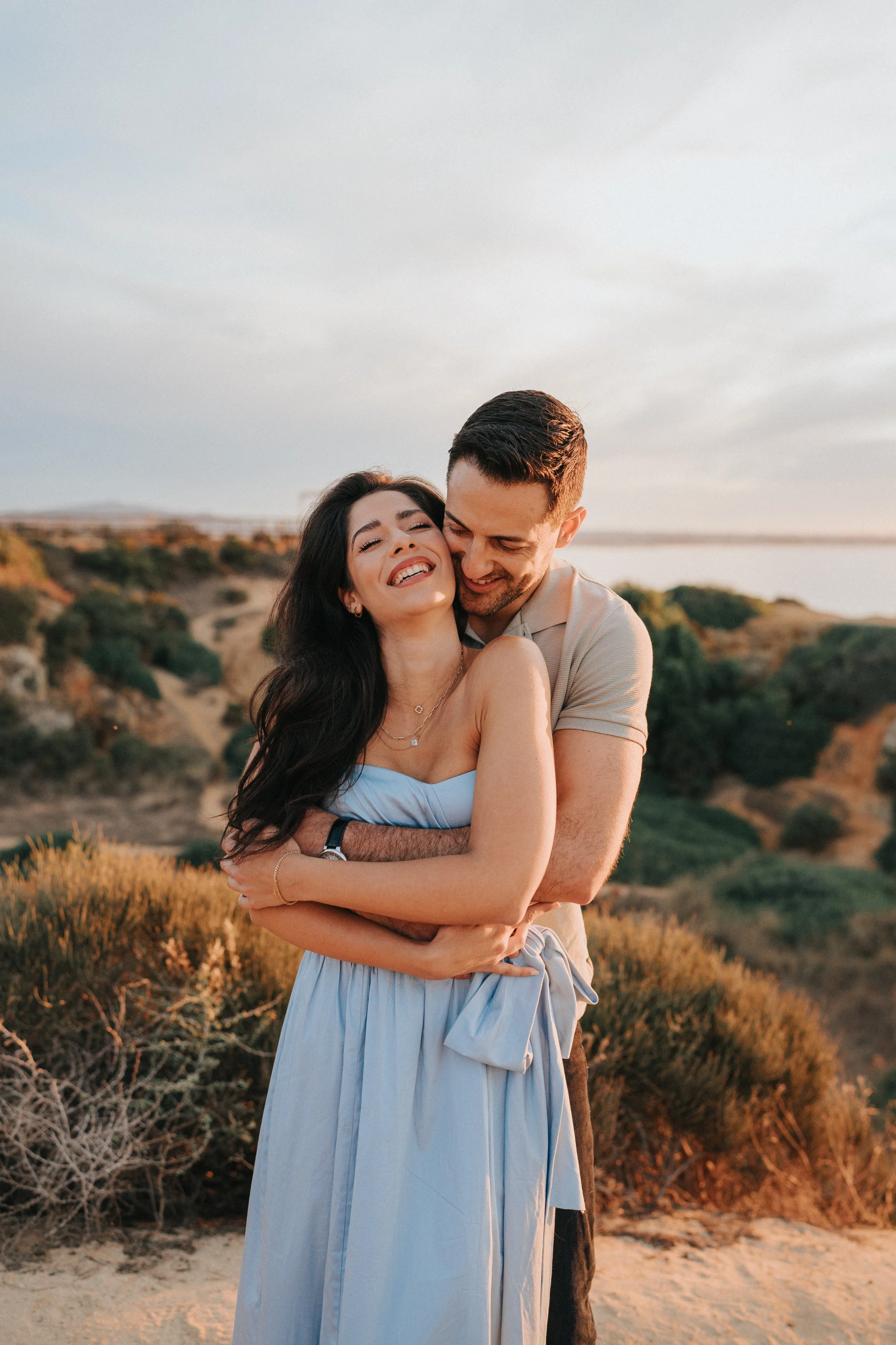 A couple embracing outdoors during sunset, with hills and vegetation in the background.