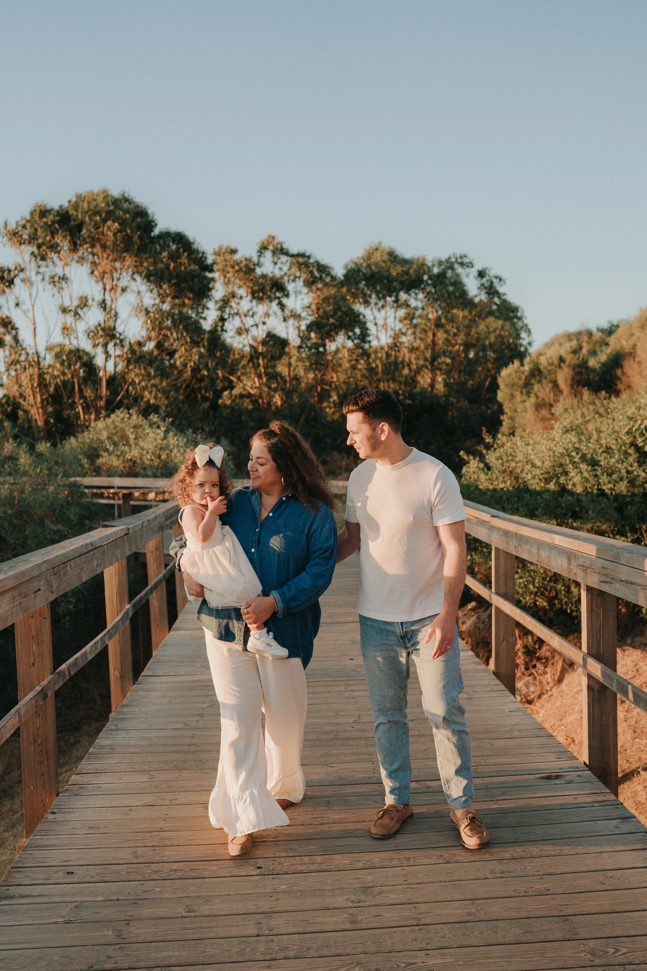 A family of three walking on a wooden bridge outdoors during sunset. The woman is carrying a young girl, and they are smiling and enjoying the moment. The man is walking beside them, looking at the girl. In the background, there are trees and a clear