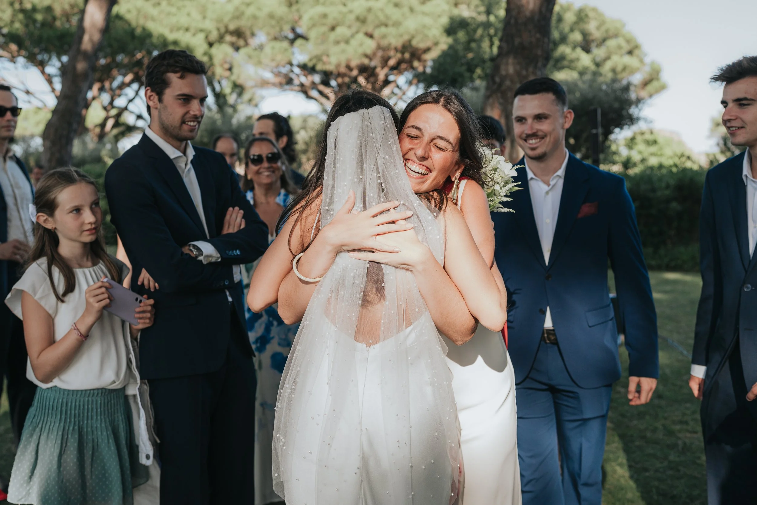 A woman in a white wedding dress is hugging a young girl wearing a pearl-embellished veil. They are surrounded by several well-dressed people outdoors with trees in the background, on a sunny day.
