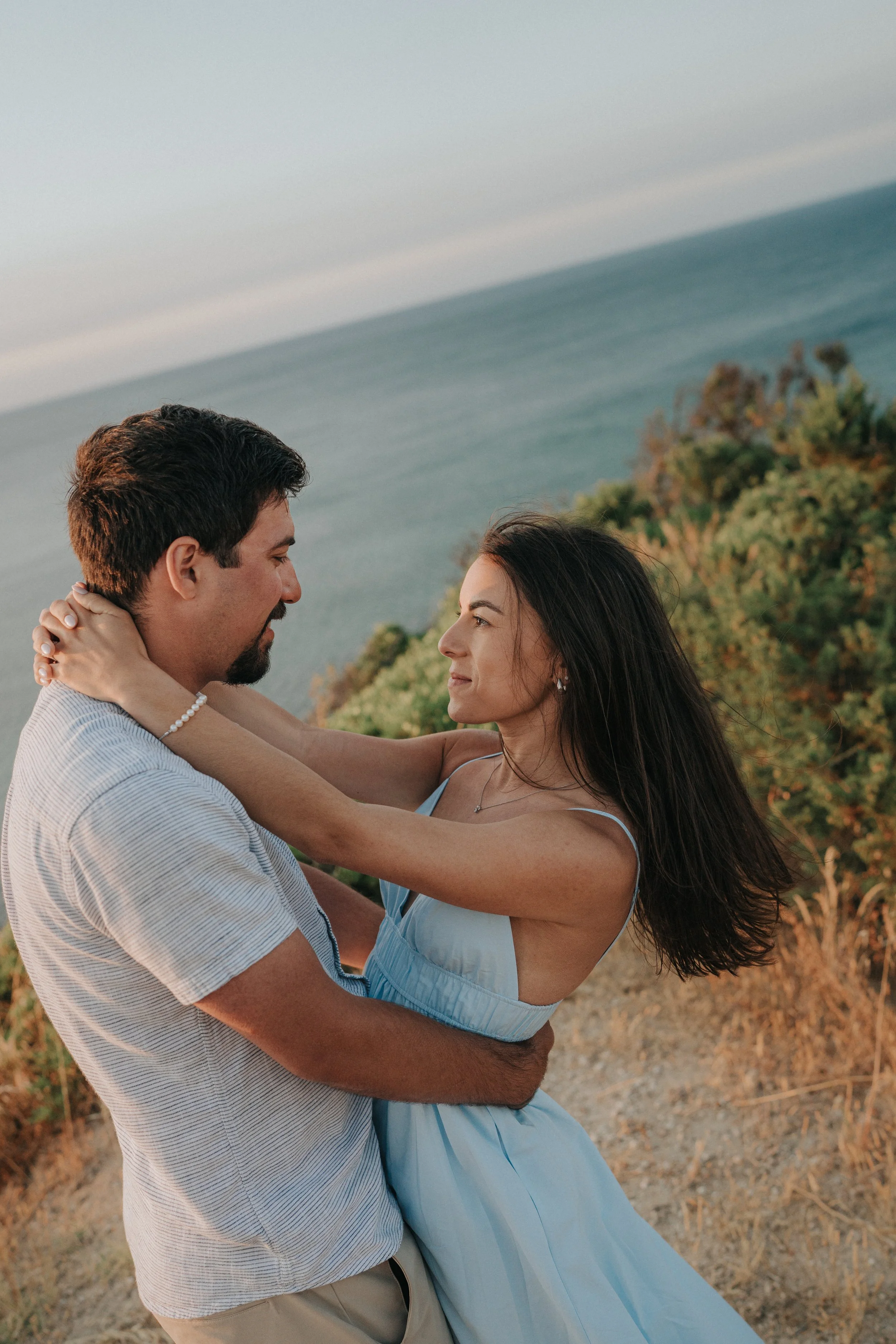 A man and a woman dance together outdoors on a hillside near the ocean, with trees and a cloudy sky in the background, both smiling and gazing into each other's eyes.