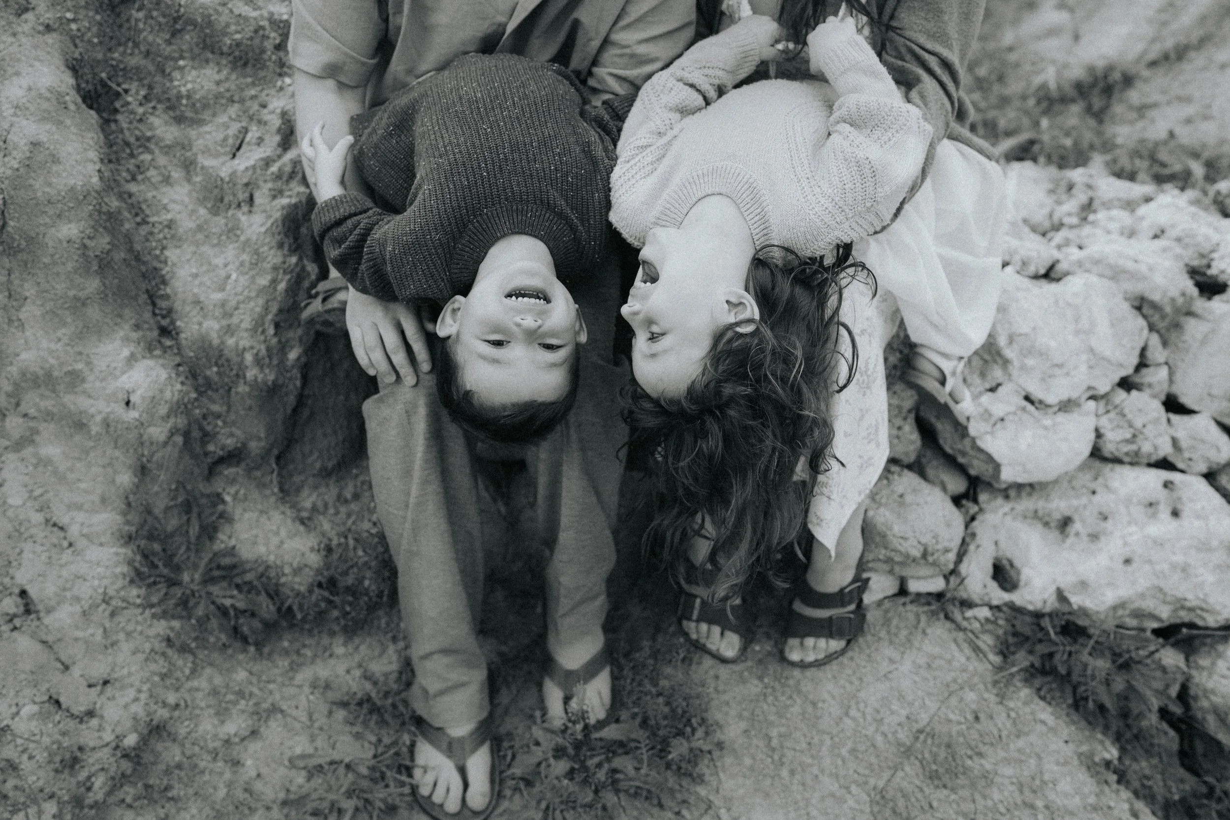 Two children with dark hair lying on rocky ground, laughing and playing together.