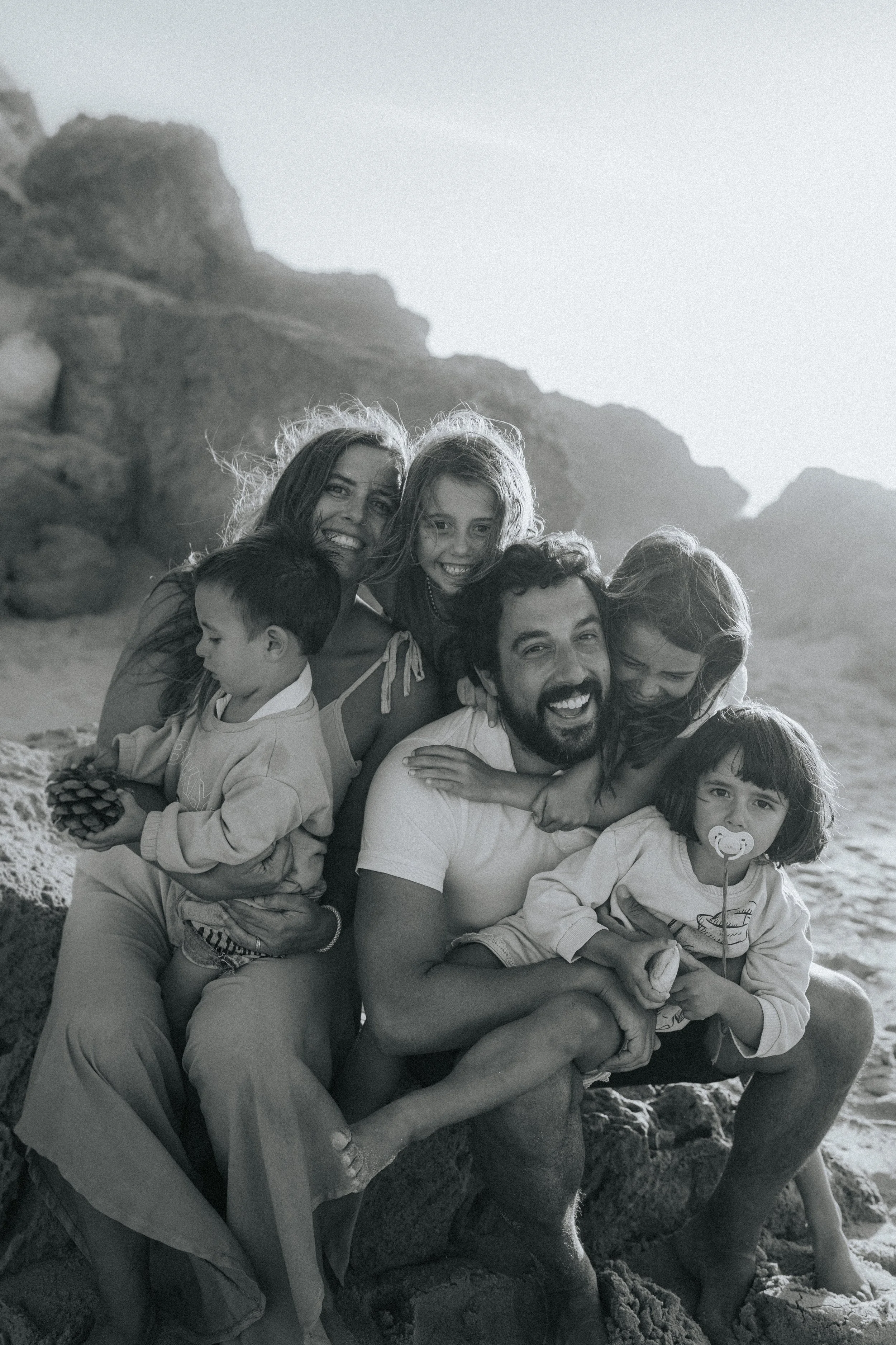 A black and white photo of a large group of people, including children and adults, smiling and hugging on a rocky beach with cliffs in the background.