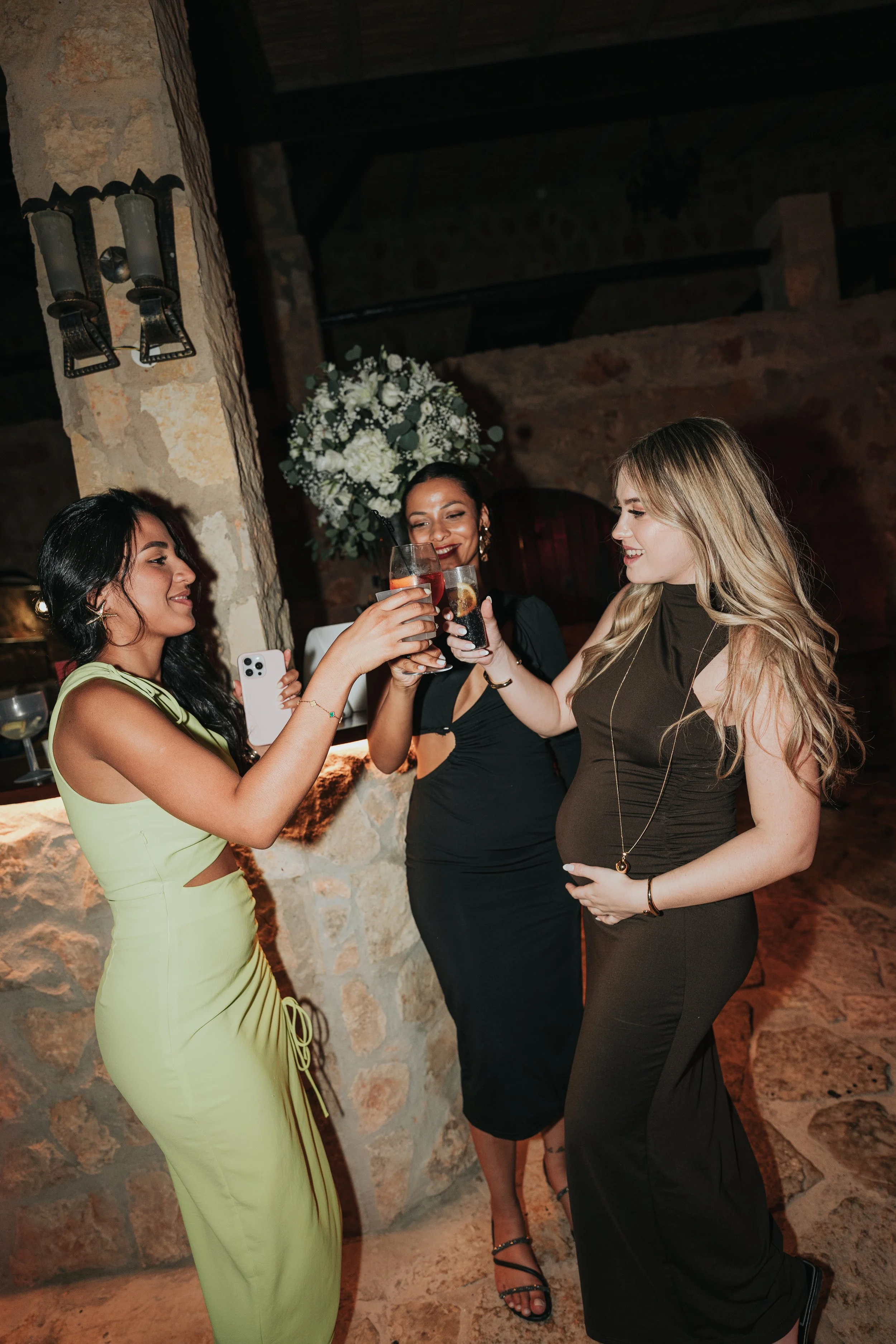 Three women celebrating with cocktails in a dimly lit rustic indoor setting, standing near a stone wall and a large floral arrangement.