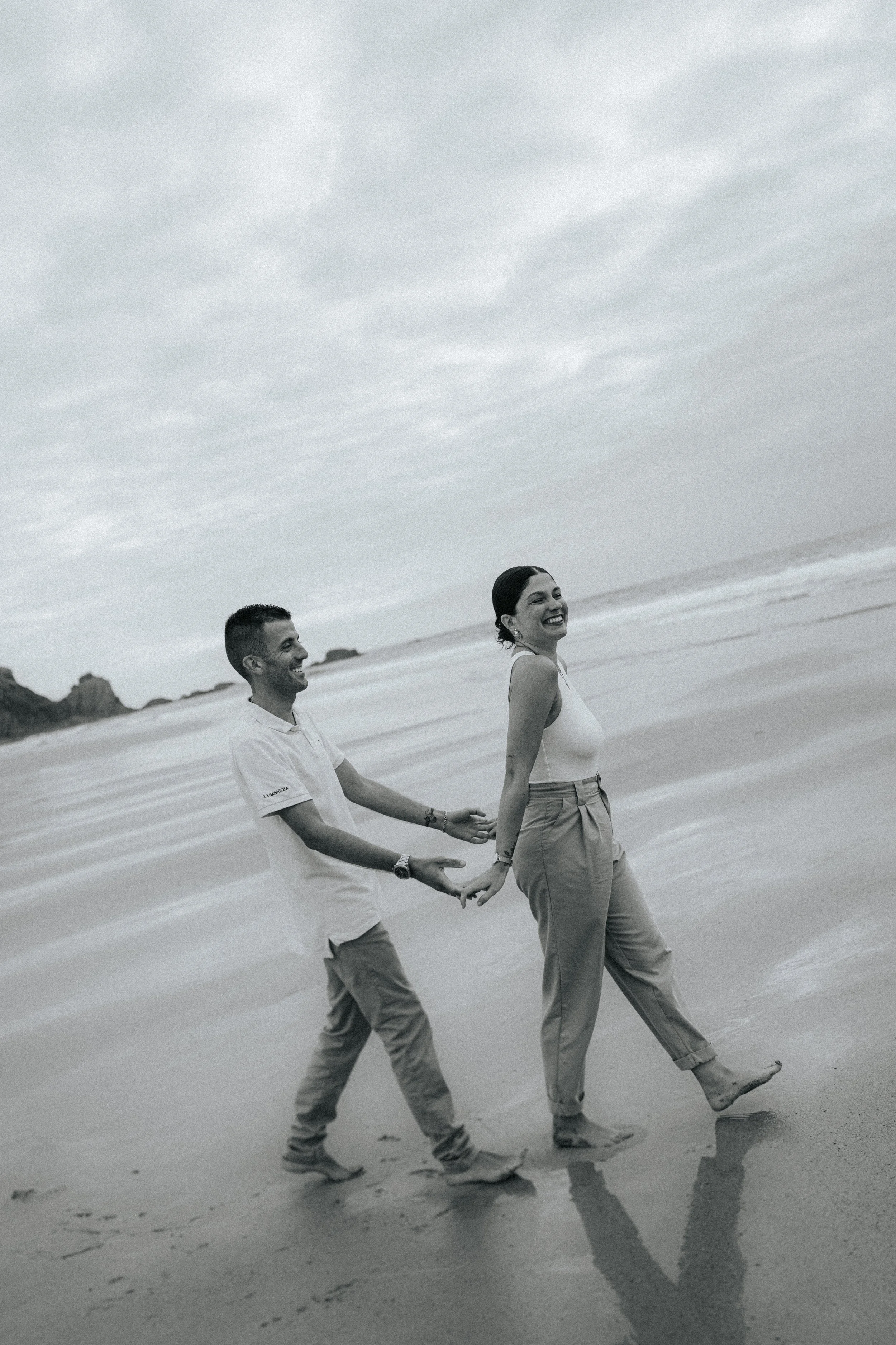 A black and white photo of a smiling woman and a man holding hands, walking barefoot on the beach, with ocean waves and cloudy sky in the background.