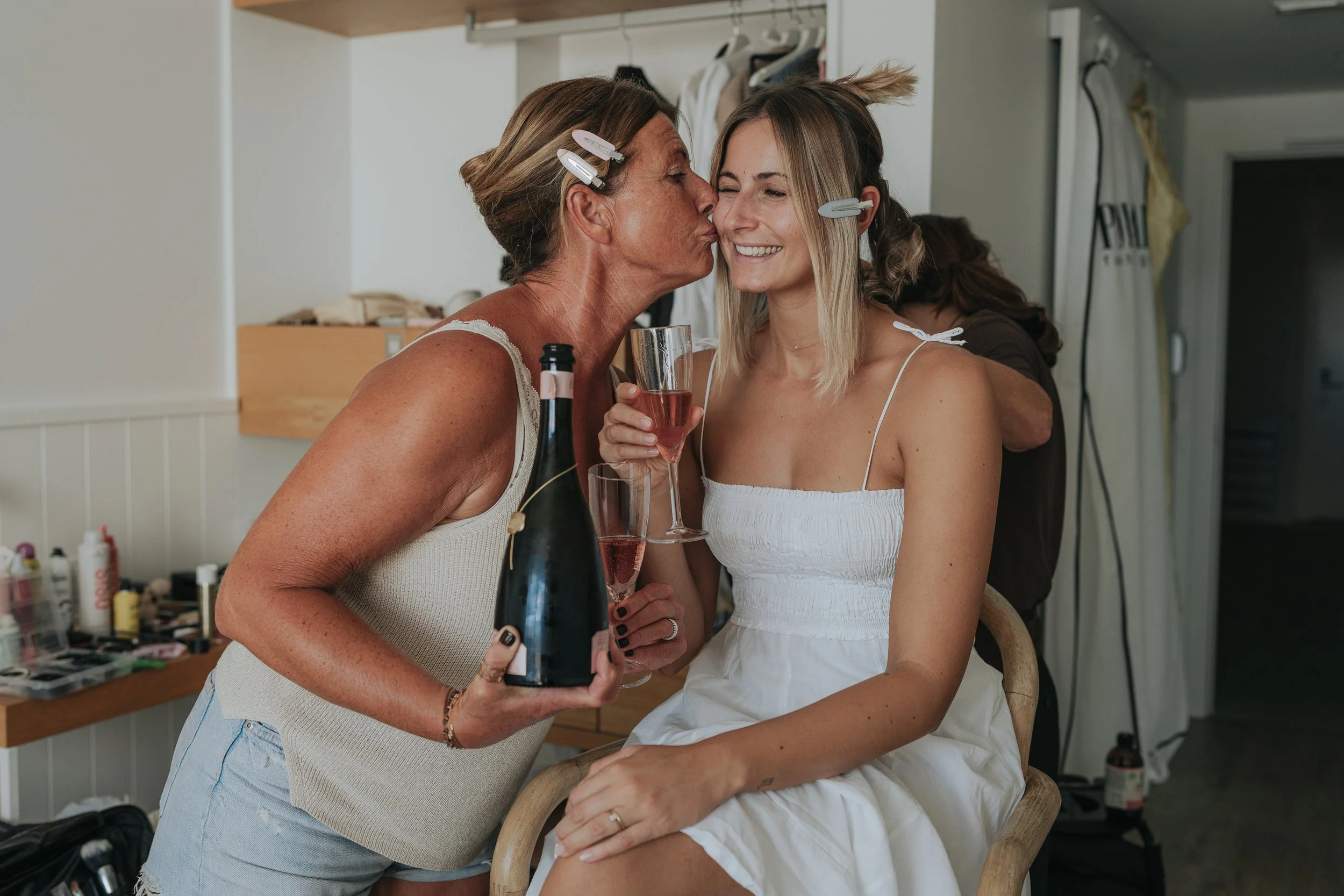 Two women are celebrating at a party, one is kissing the other's cheek while holding a bottle of champagne and a glass of pink wine. The woman getting kissed is smiling and wearing a white dress. Both women are wearing hair clips.
