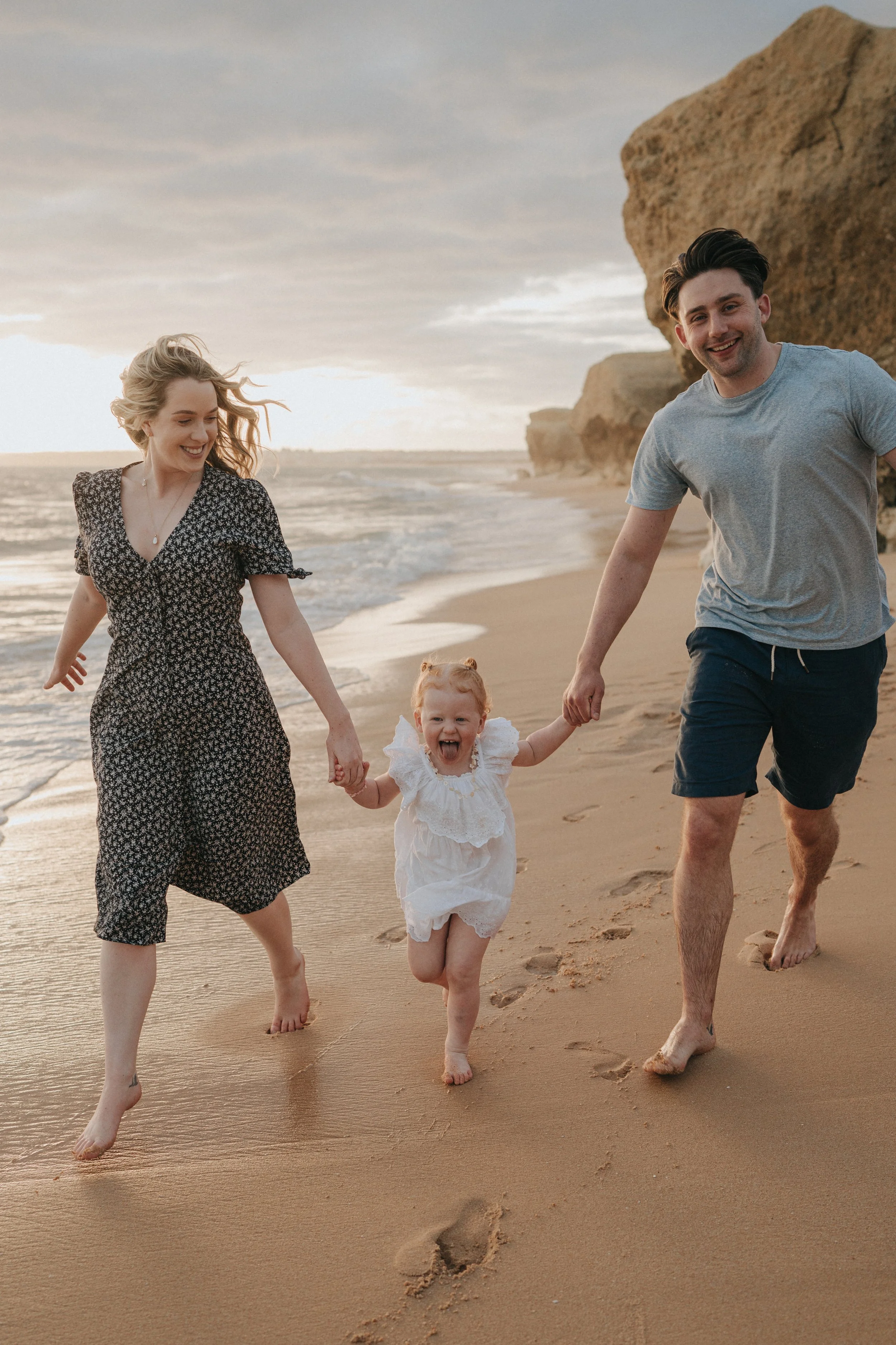 A family of four—two adults and two children—are walking barefoot on the beach near the shoreline, with rocky cliffs in the background during sunset, smiling and holding hands.