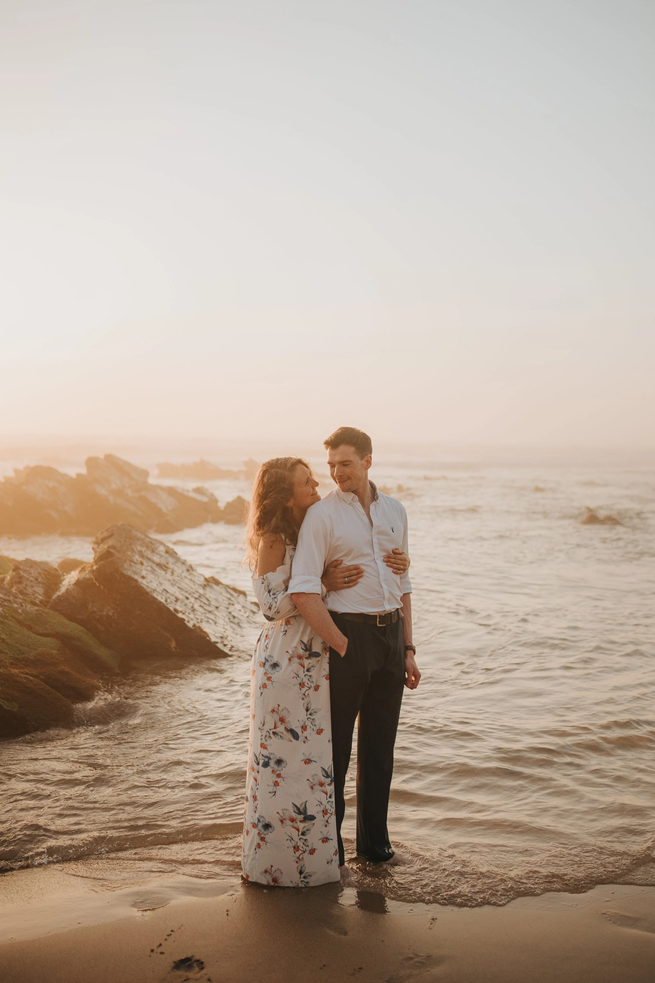 A couple embracing at the beach during sunset, with rocky shoreline and ocean waves in the background.