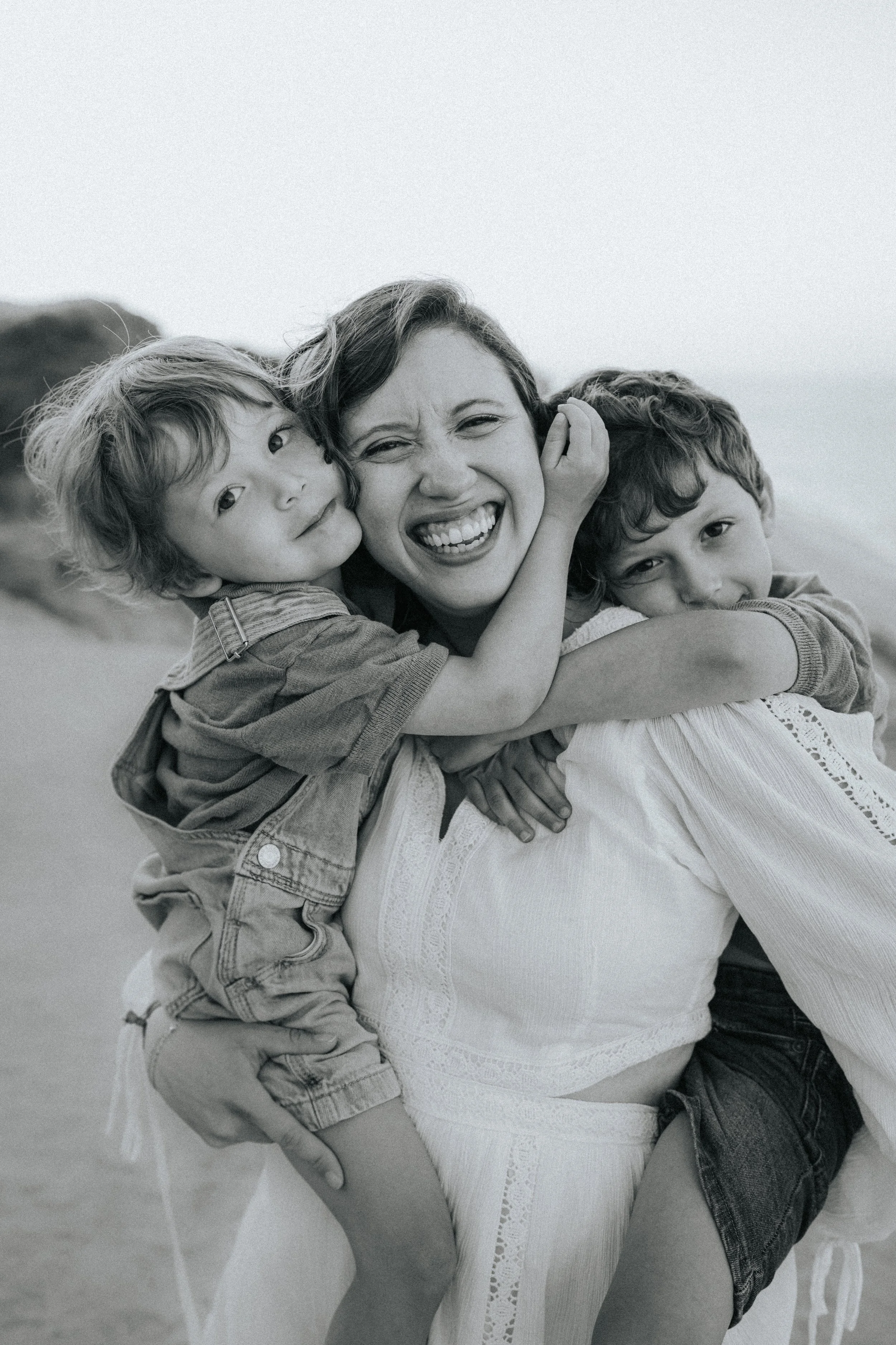 Black and white photo of a woman with two children, all smiling and hugging each other outdoors.
