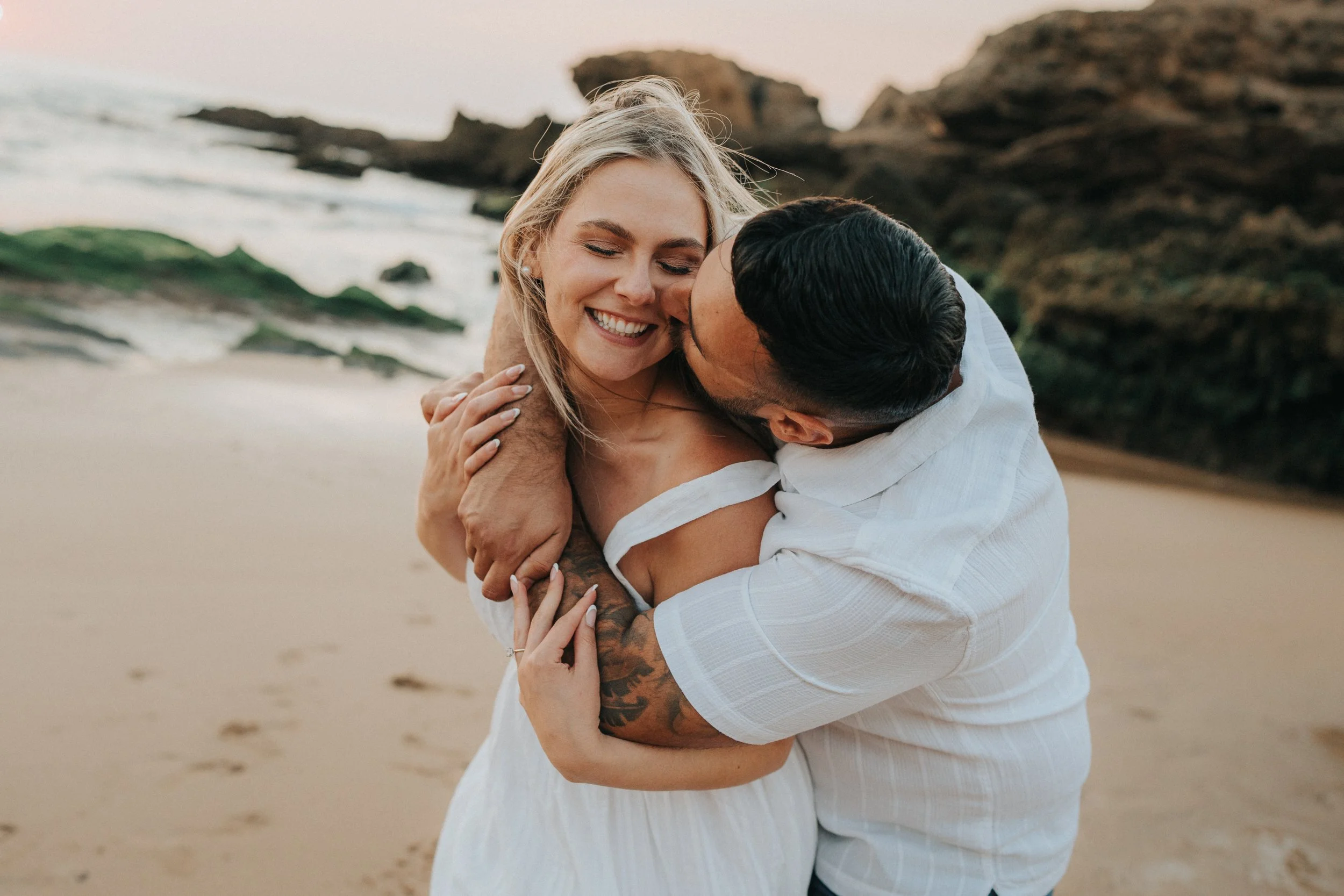 A happy couple hugging on a beach with rocks and the ocean in the background at sunset.
