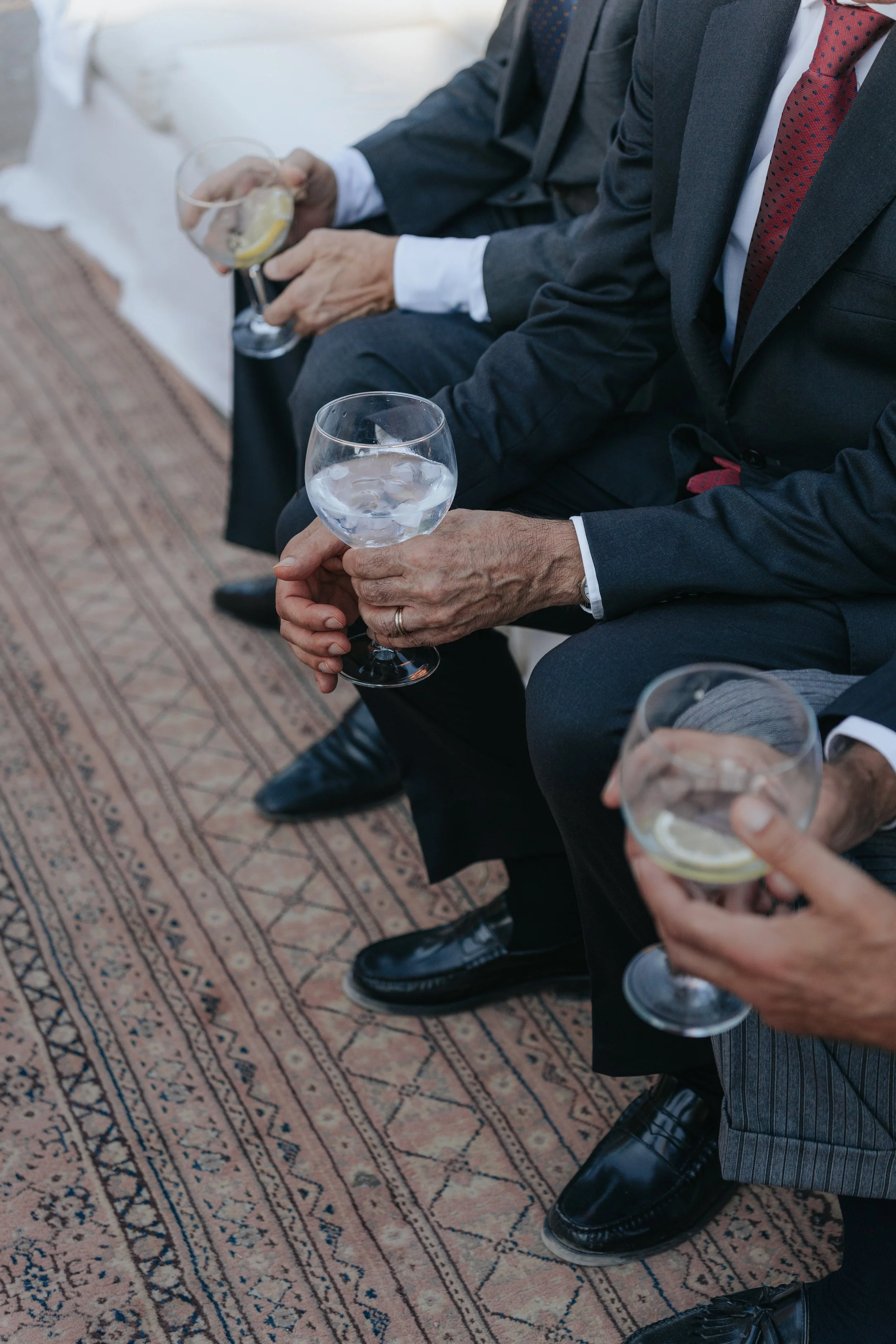 Close-up of several men's hands holding drinking glasses with clear liquid, ice, and lemon slices, sitting on a patterned carpet in formal suits.