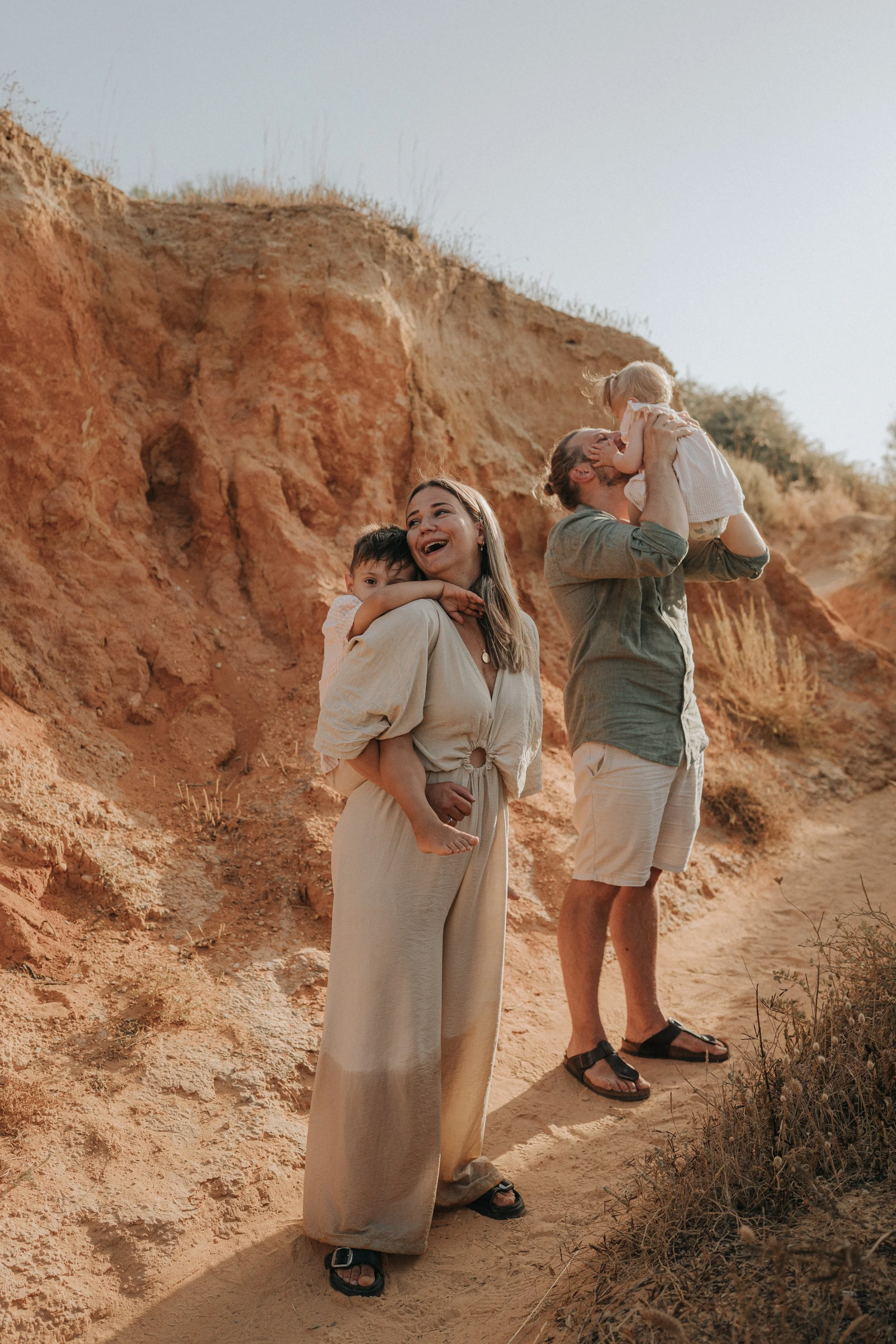 A family of four enjoying a sunny day outdoors near a dirt hill. The mother stands holding a child on her back, smiling and wearing a beige dress. The father is lifting a young girl, kissing her on the face, and wearing a green shirt and shorts. The 