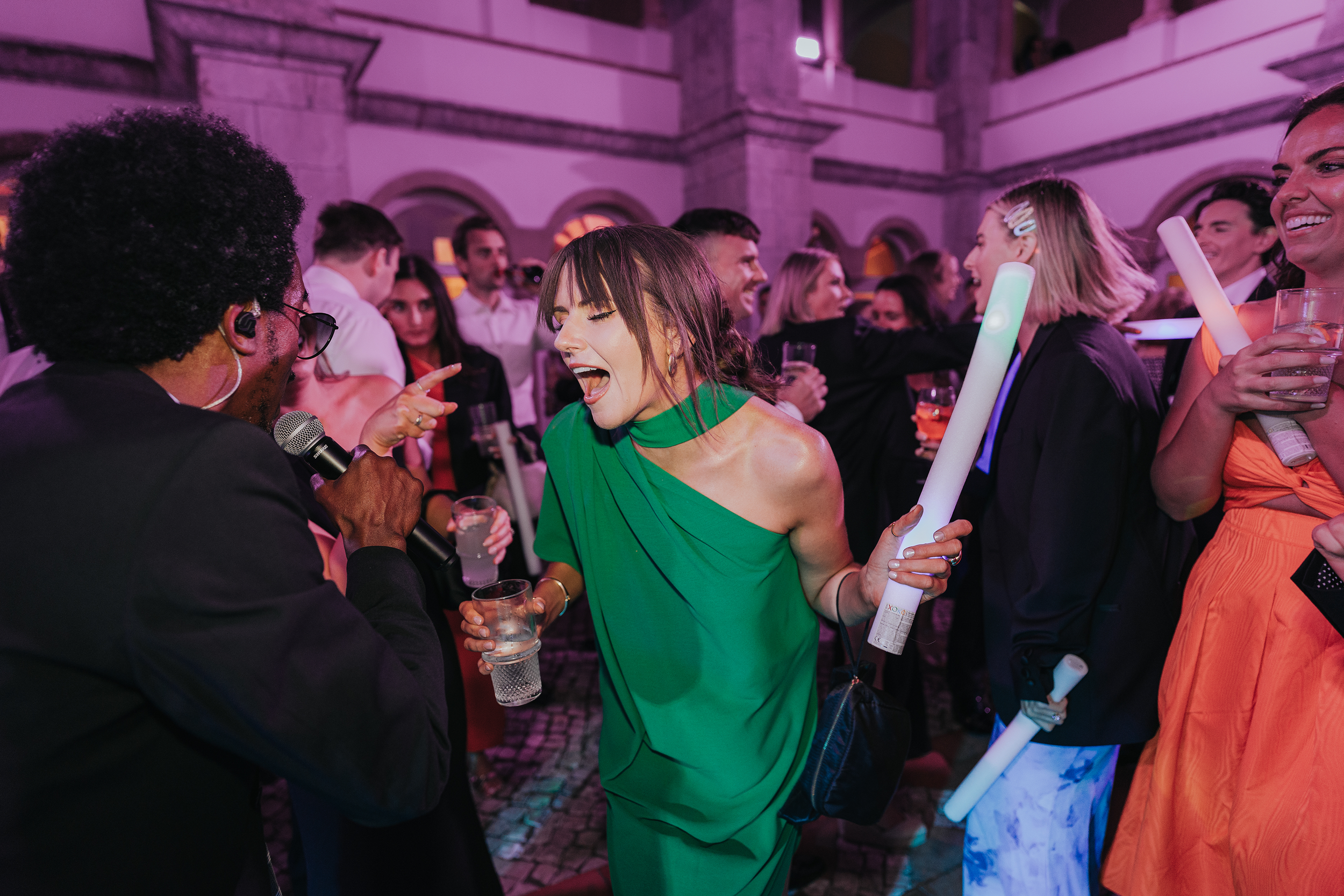 A lively party scene with people dancing and socializing. A woman in a green dress is smiling and holding a glass of water, while a man with an afro hairstyle and glasses is speaking into a microphone. The atmosphere is vibrant with colorful lighting