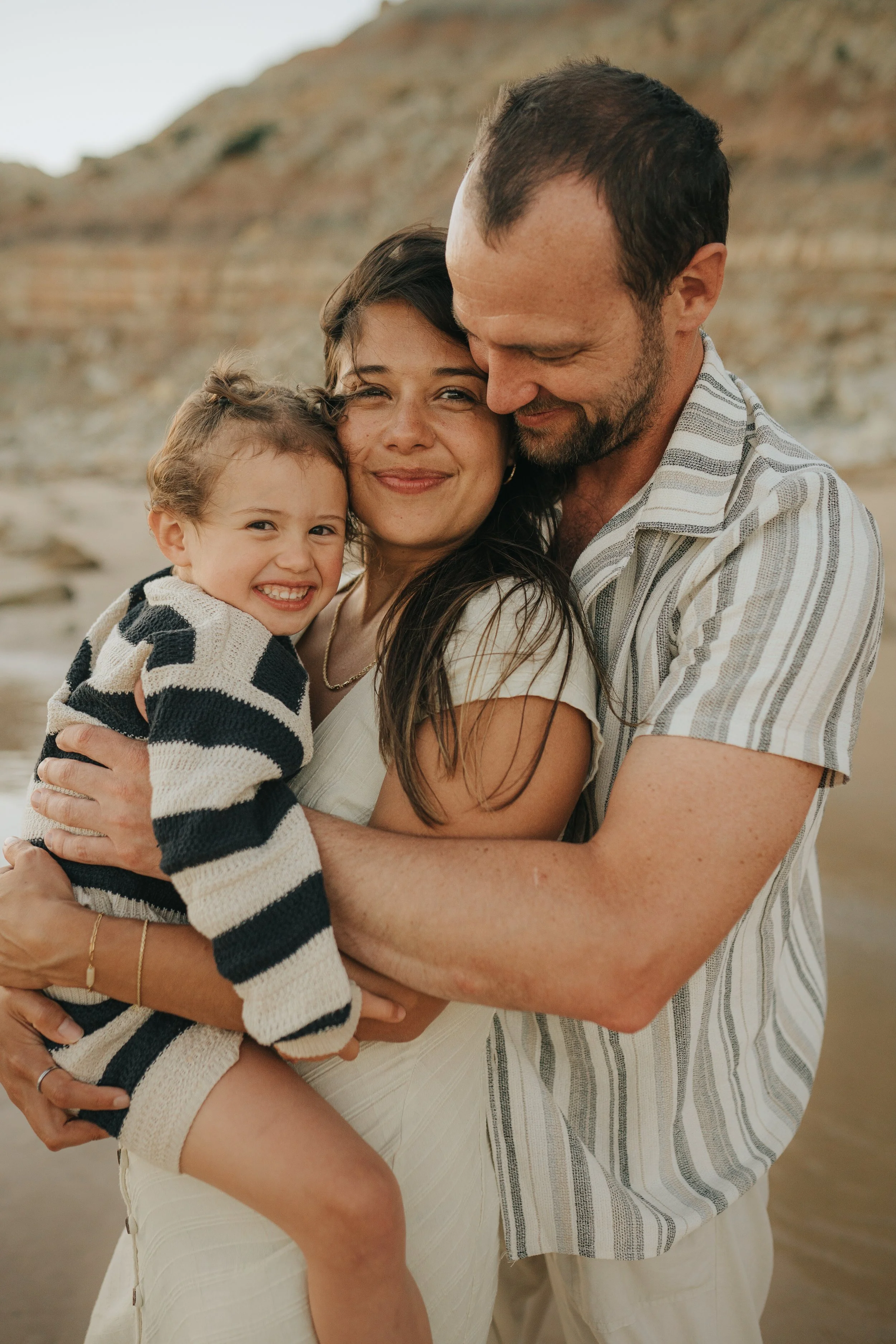 A family of three hugging on a beach with rocky cliffs in the background. The mother has dark hair, and the father has short dark hair and a beard. The daughter has curly hair and is smiling widely.