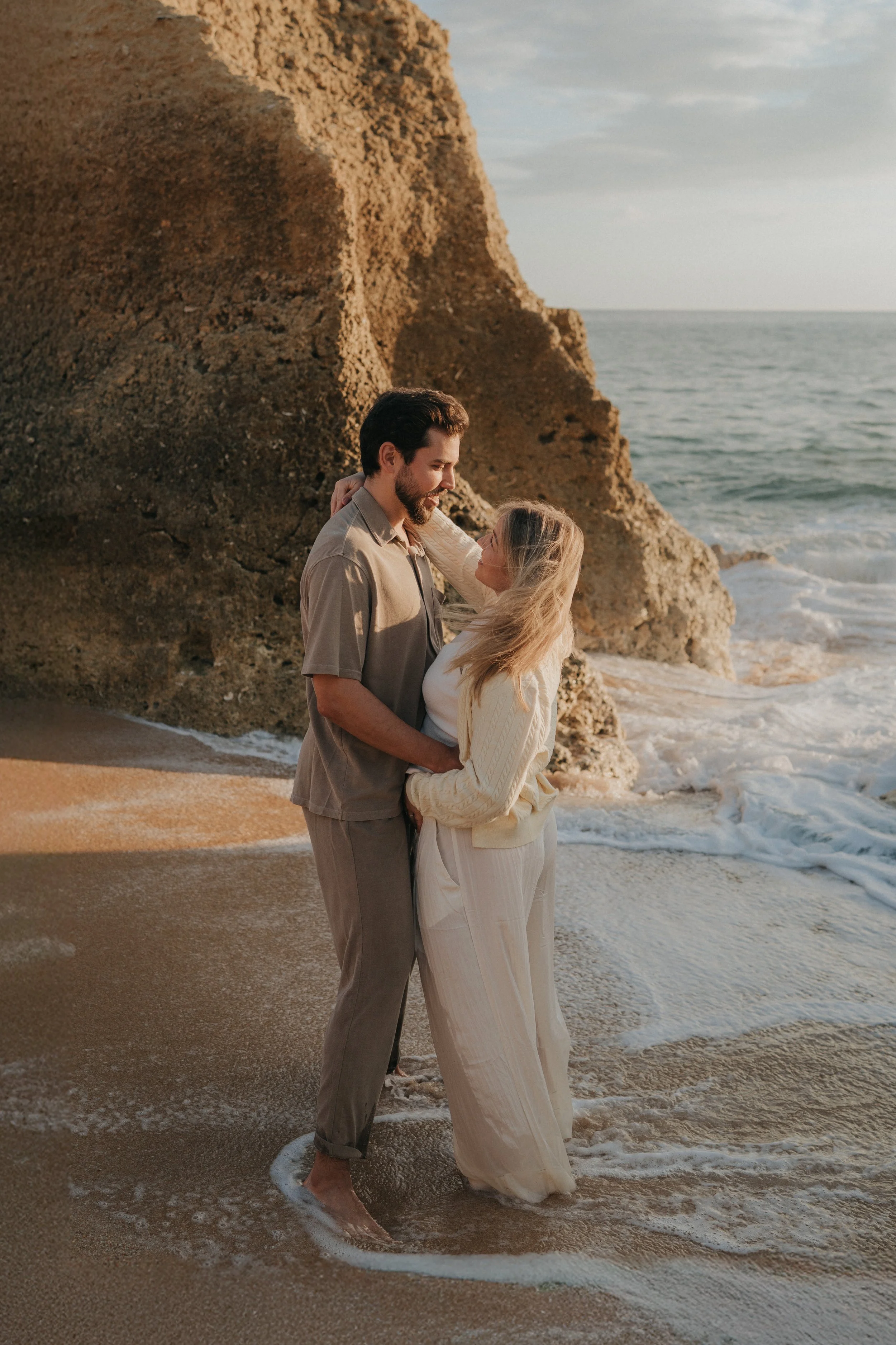 A couple embracing in shallow ocean water near rocky cliffs at sunset, with the woman in light-colored clothing and the man in a beige shirt and pants.