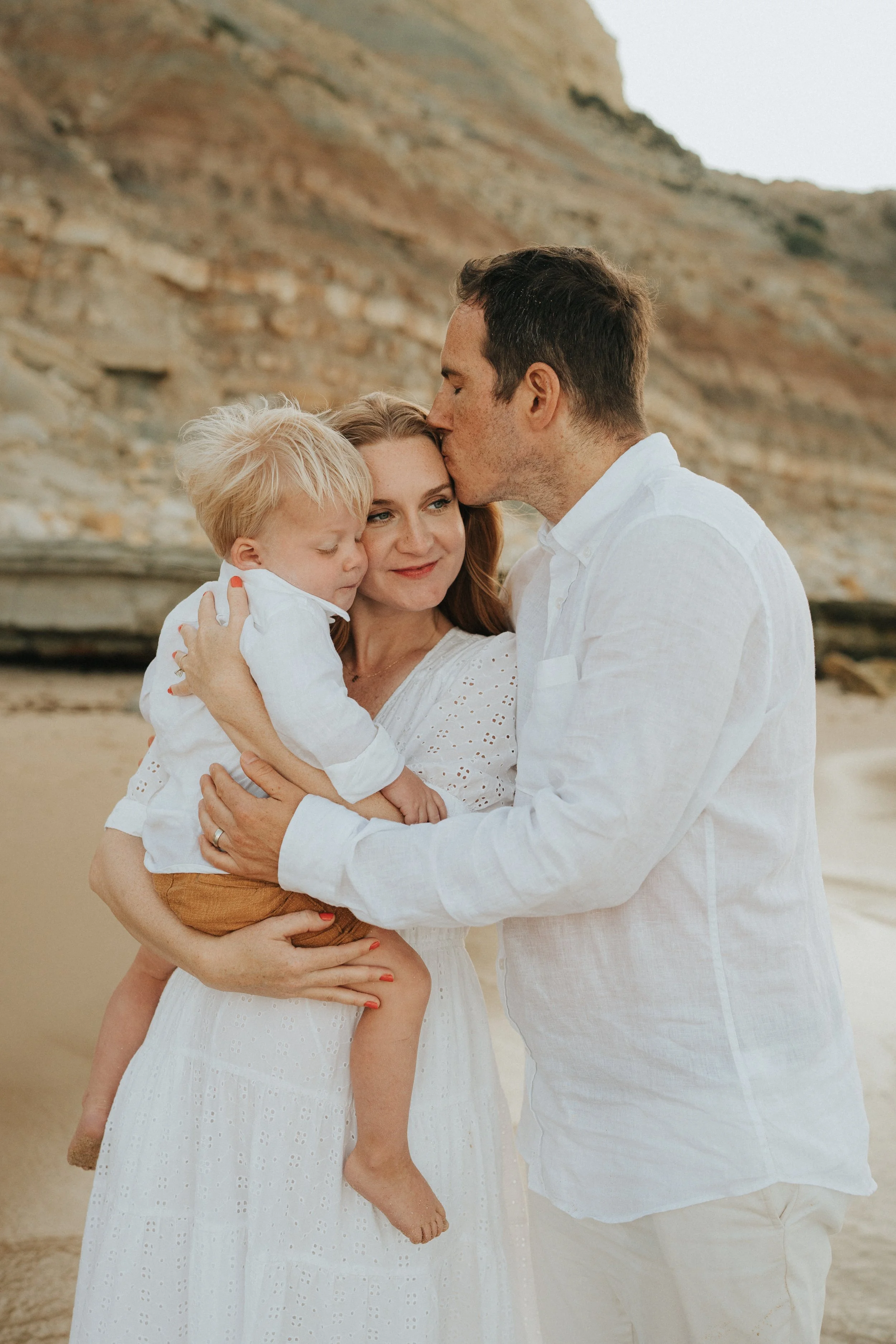 A family of three, a father, mother, and young son, on a beach with cliffs in the background. The father is kissing the mother on the forehead while the mother holds their son warmly. The mother wears a white dress, the son has a white shirt and brow