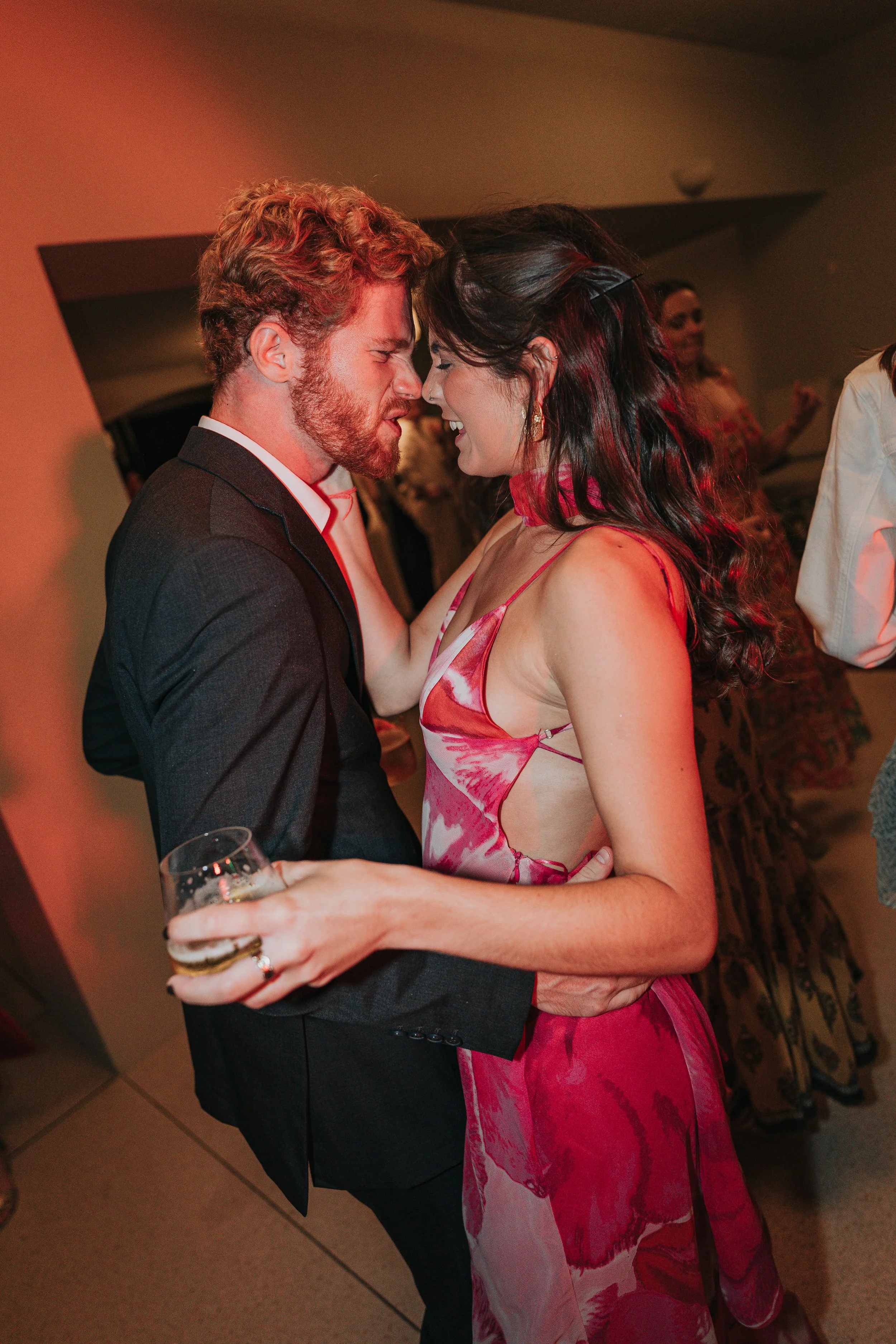 A couple dancing closely at a social event. The man has curly red hair and a beard, wearing a black tuxedo. The woman has long dark hair with red highlights, wearing a pink and white patterned dress. They are smiling and holding a glass of champagne.