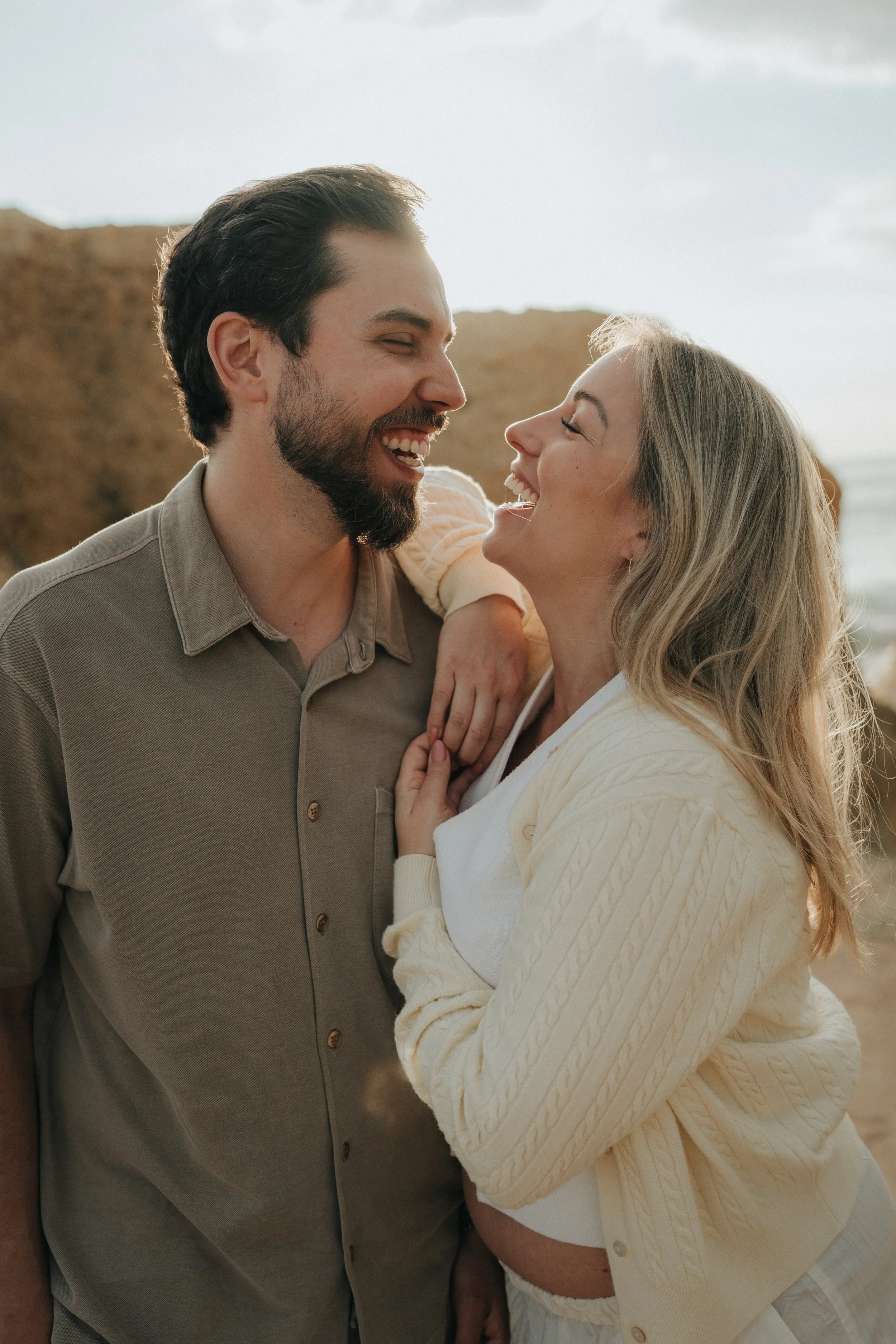 A happy couple smiling and laughing together at the beach, with ocean and cloudy sky in the background. The man has dark hair and a beard, and is wearing a beige button-up shirt. The woman has blonde hair and is wearing a cream-colored knitted cardig