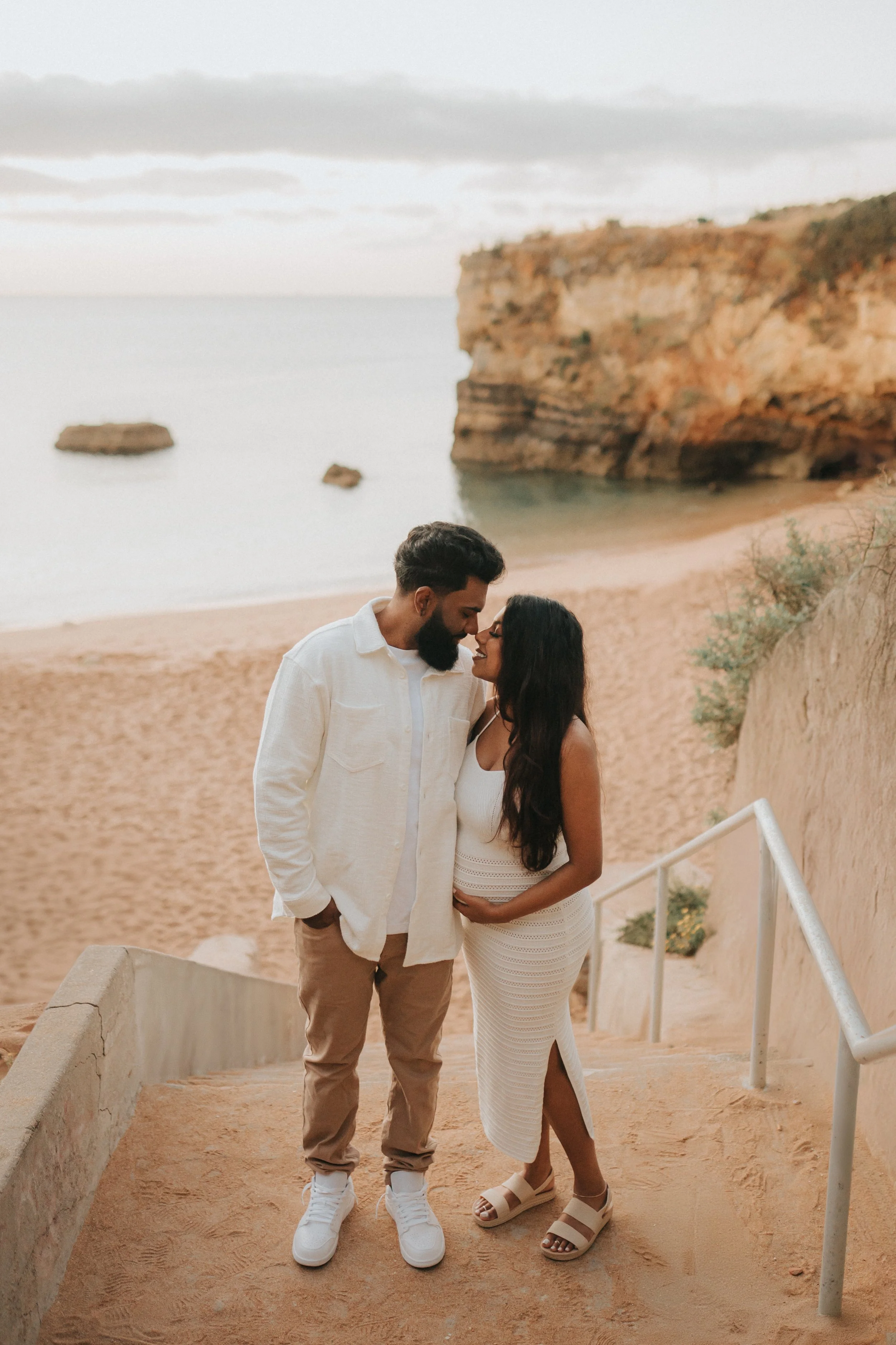 A couple standing on a pathway on a beach with cliffs in the background, looking at each other lovingly.