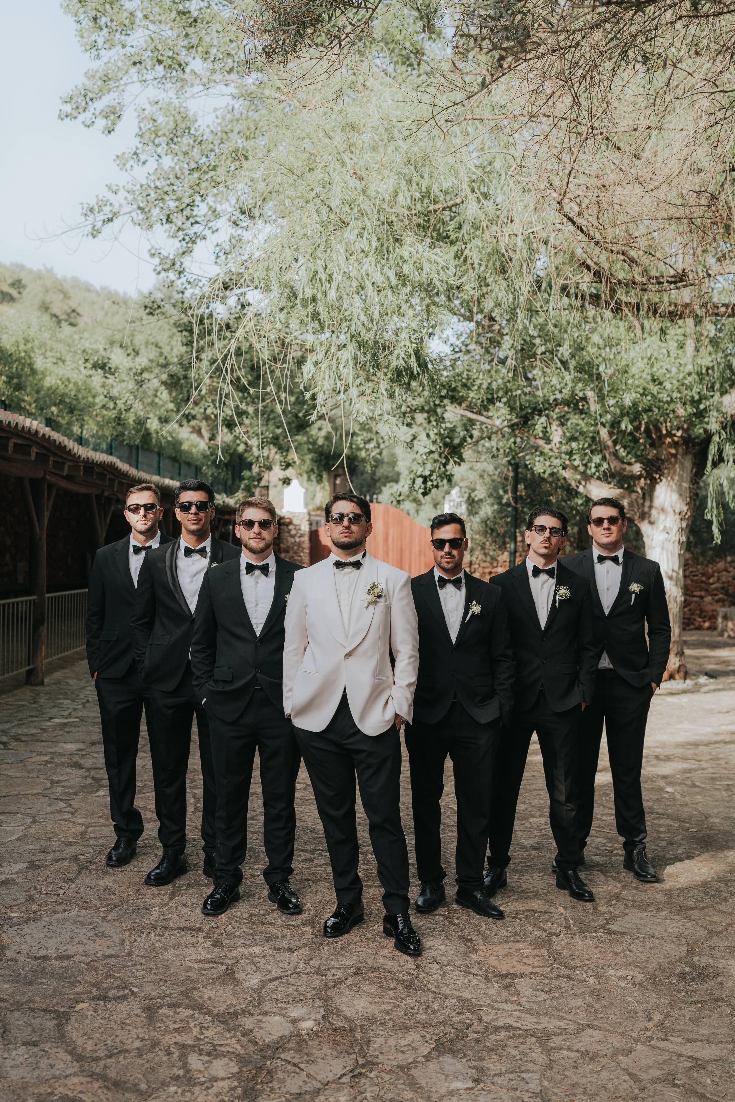 A group of nine men in tuxedos walking outdoors on a cobblestone path beneath trees, with some wearing sunglasses and white boutonnières.