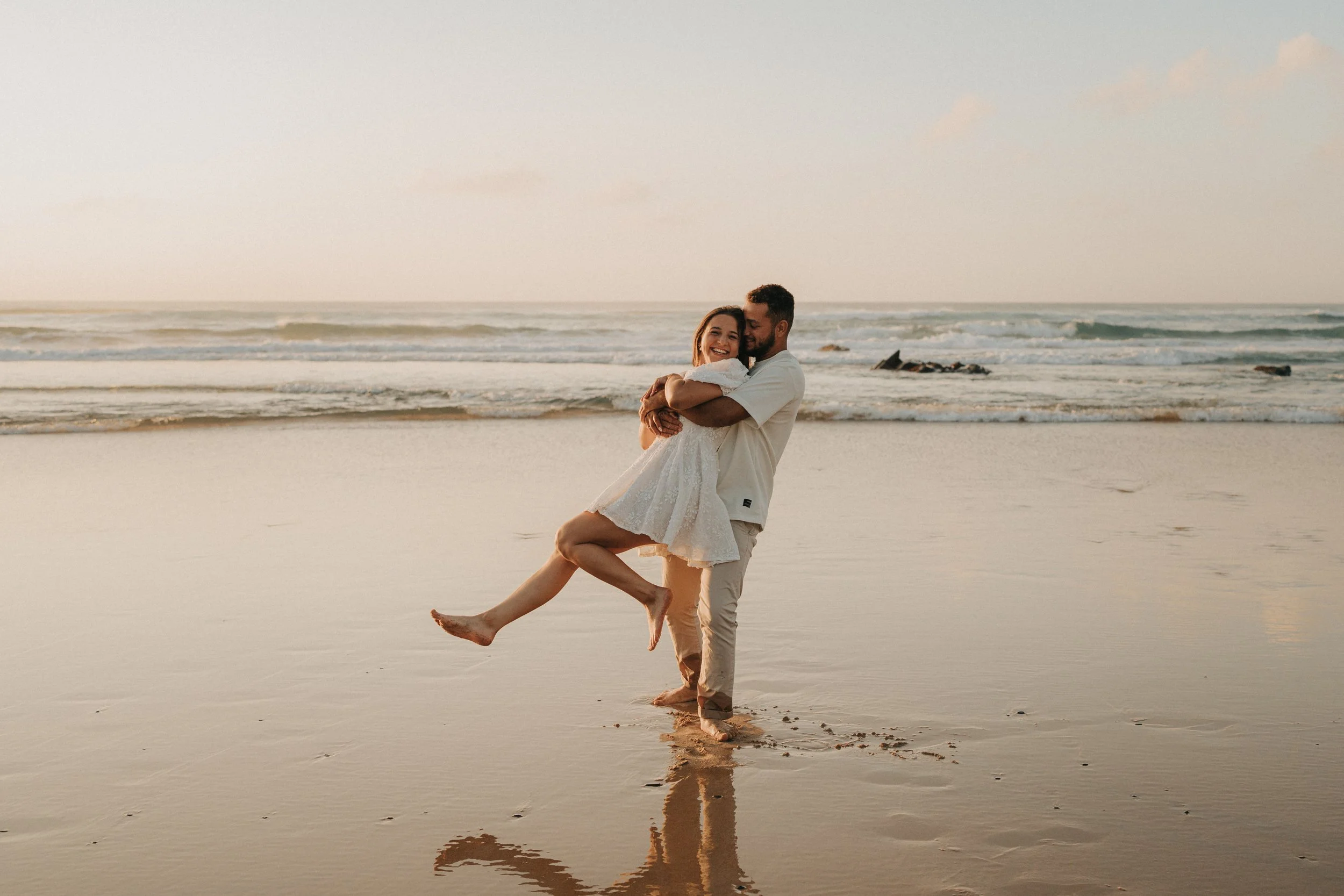 A man carrying a woman on a beach at sunset, with the ocean and waves in the background.