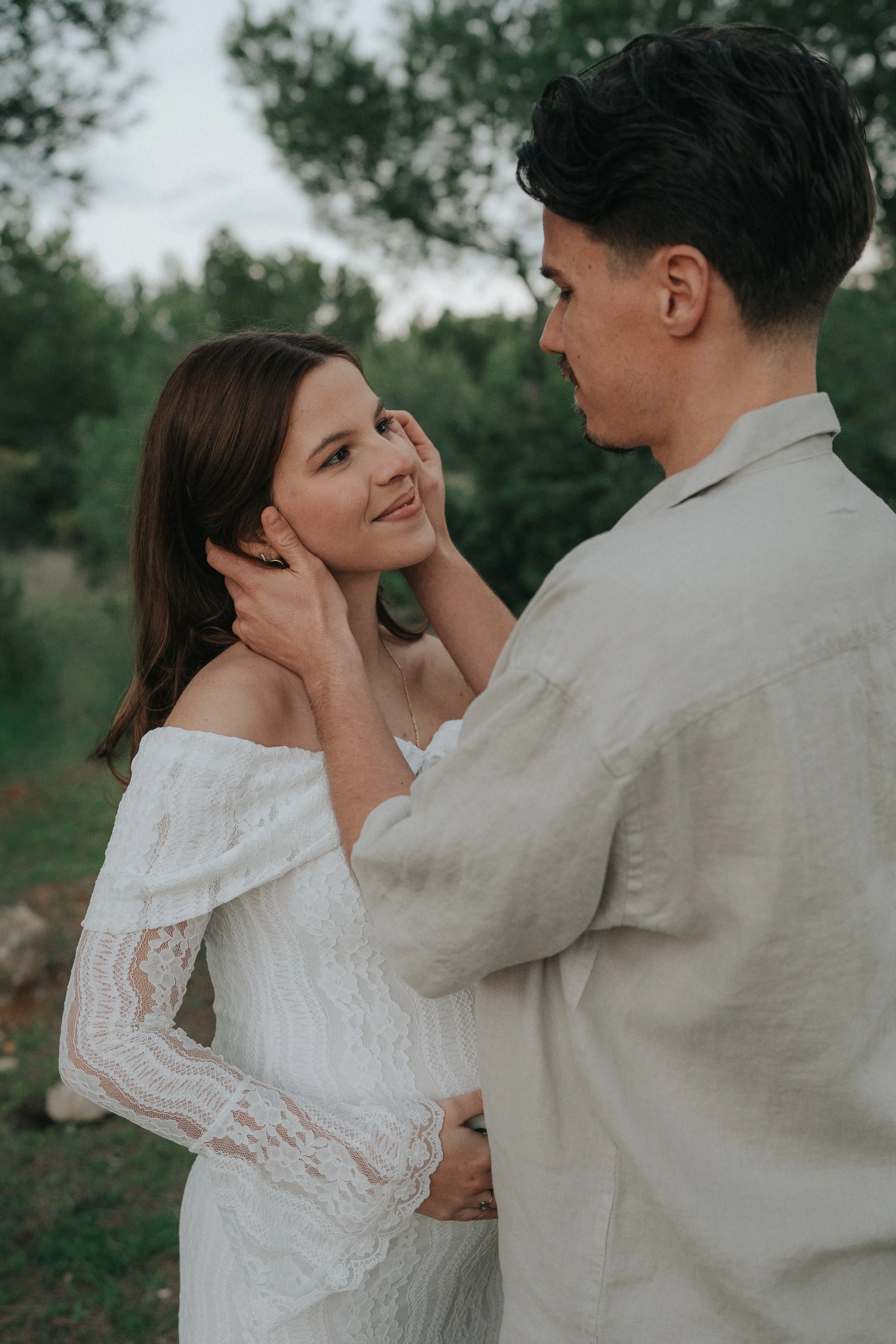 A man tenderly holds a woman's face and pregnant belly in an outdoor setting, both smiling at each other.