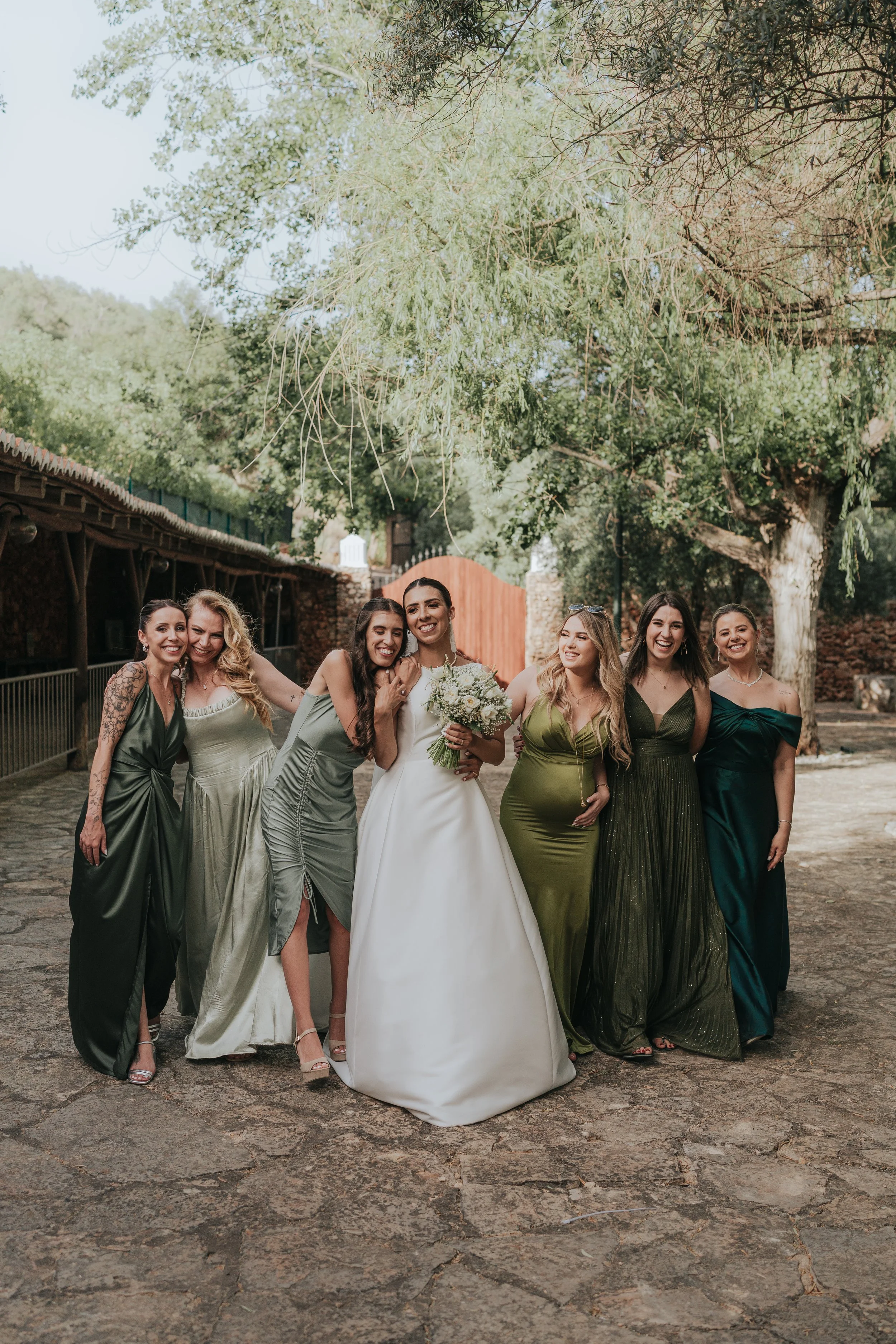 A group of seven women in formal dresses standing outdoors on a stone path, with trees and a wooden gate in the background, celebrating a wedding.