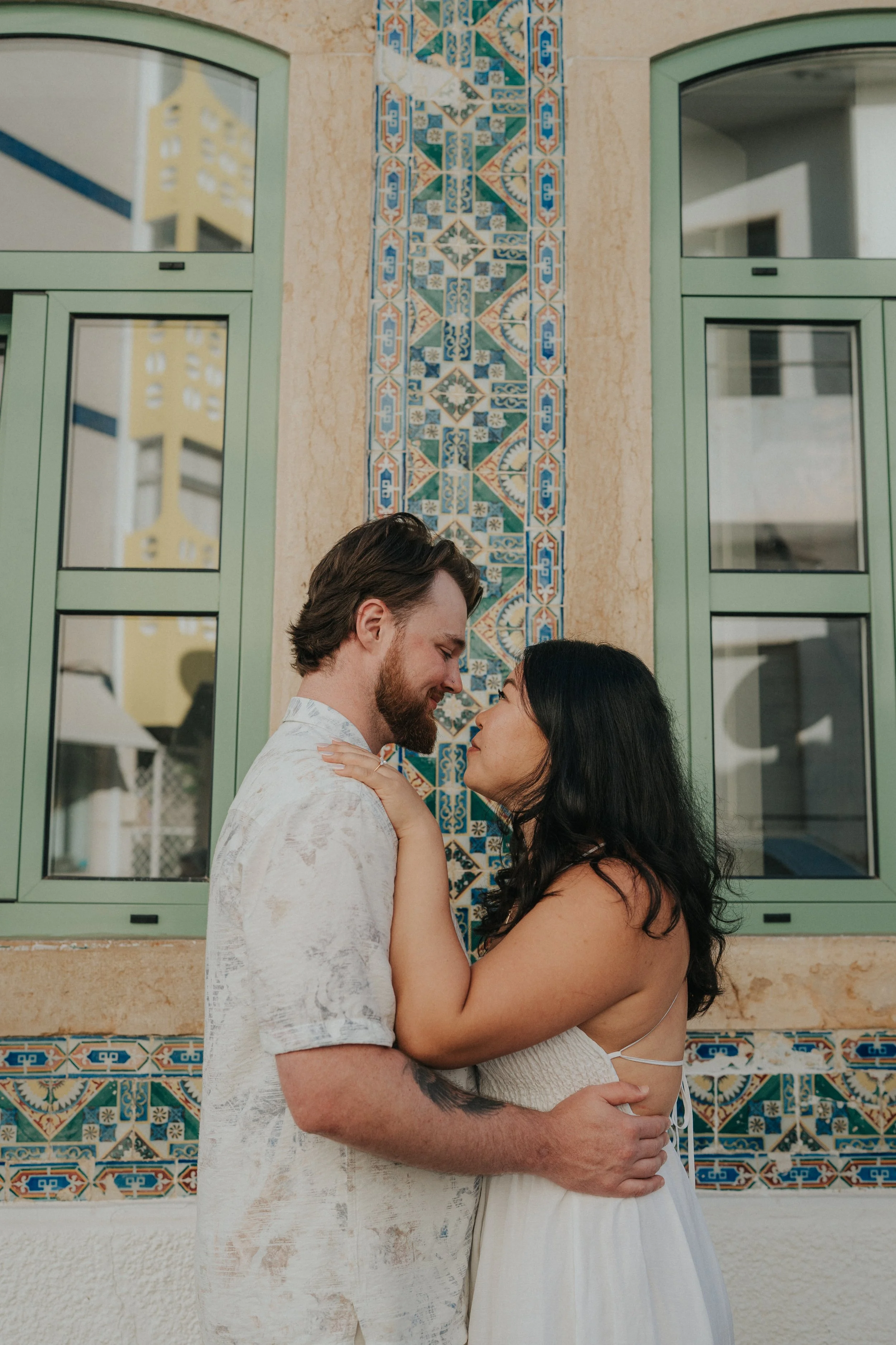 A couple embracing and looking at each other closely in front of a decorative tile wall and window.