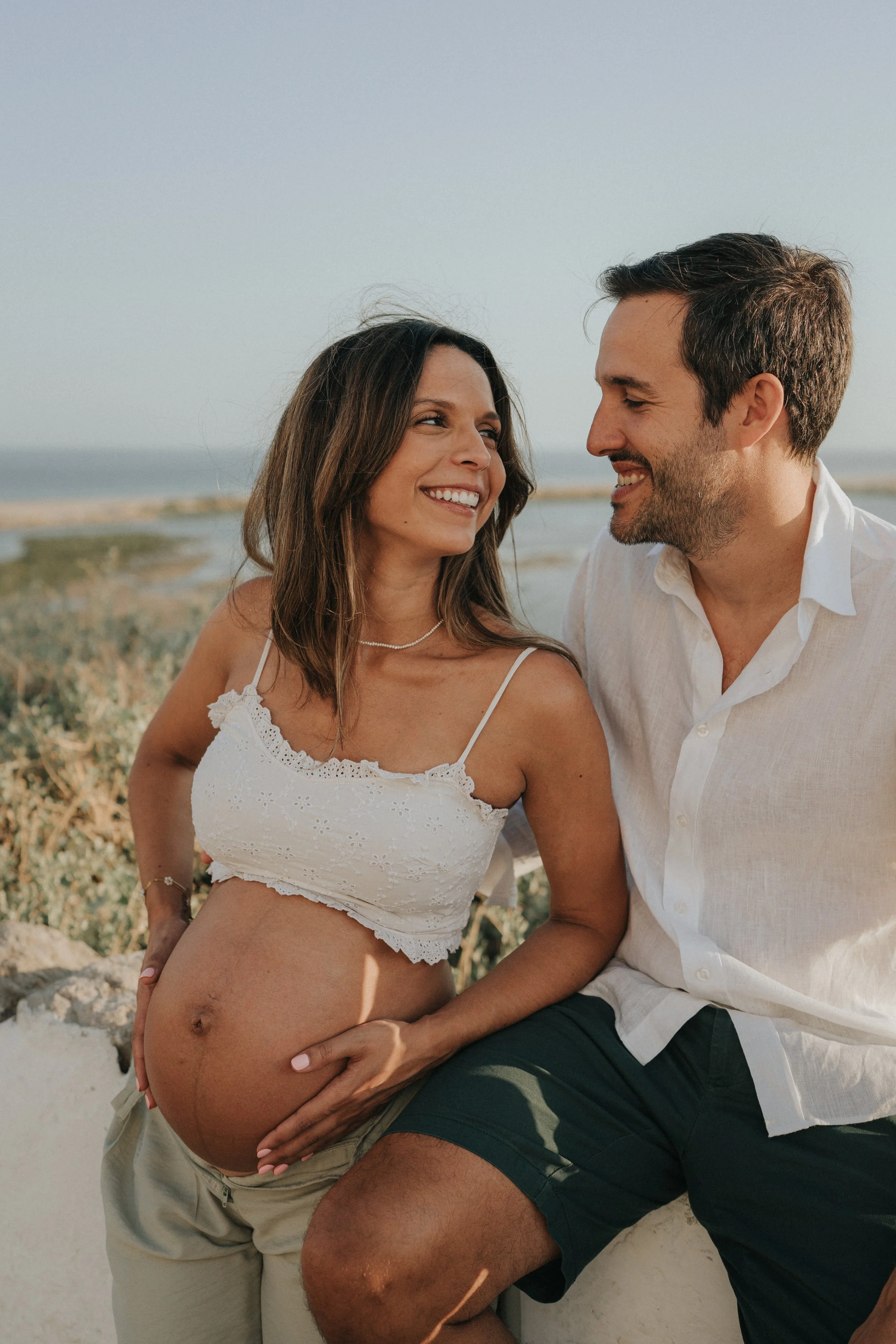 A pregnant woman in a white lace crop top sharing a joyful moment with a man in a white shirt at the beach.