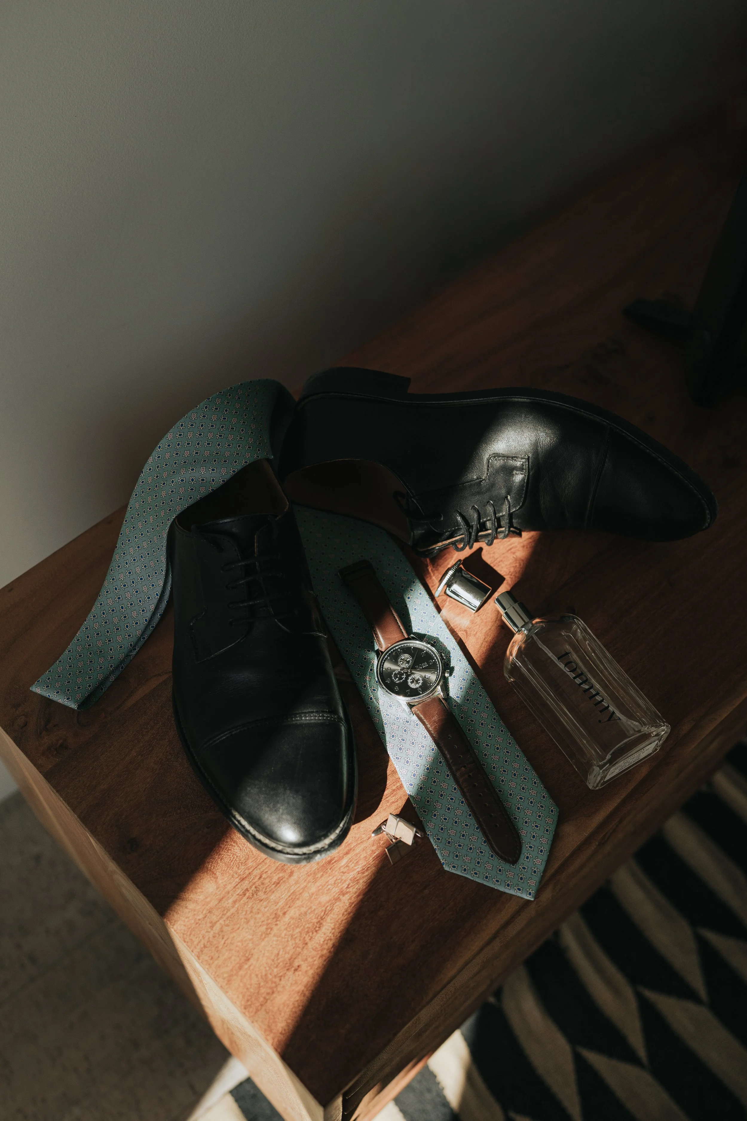 Black leather shoes, a wristwatch, a patterned tie, a brown belt, a perfume bottle, and a water bottle placed on a wooden table.