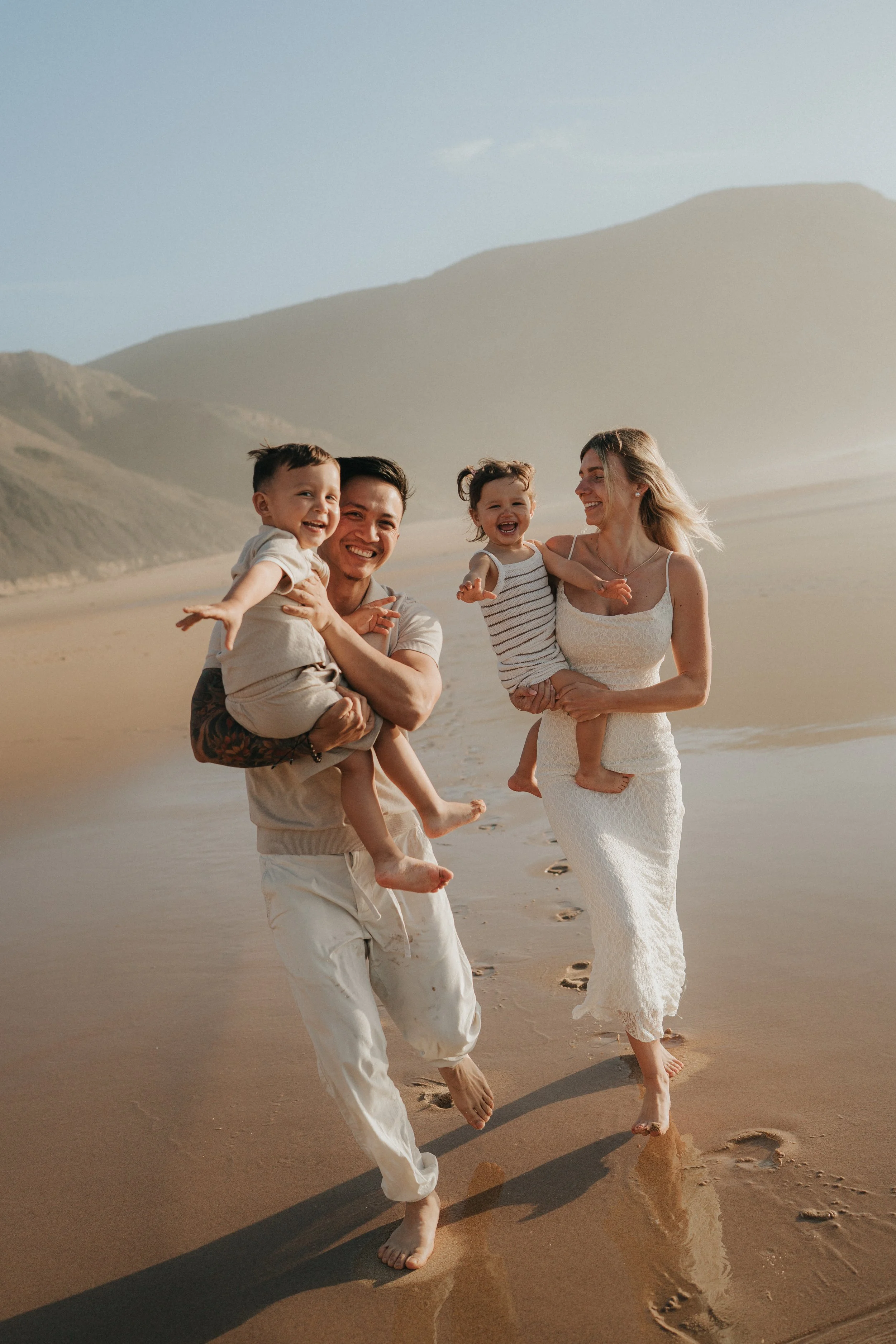 Family of four enjoying a walk on the beach, with two young children and two adults, all smiling and barefoot, with mountainous landscape in the background during sunset.
