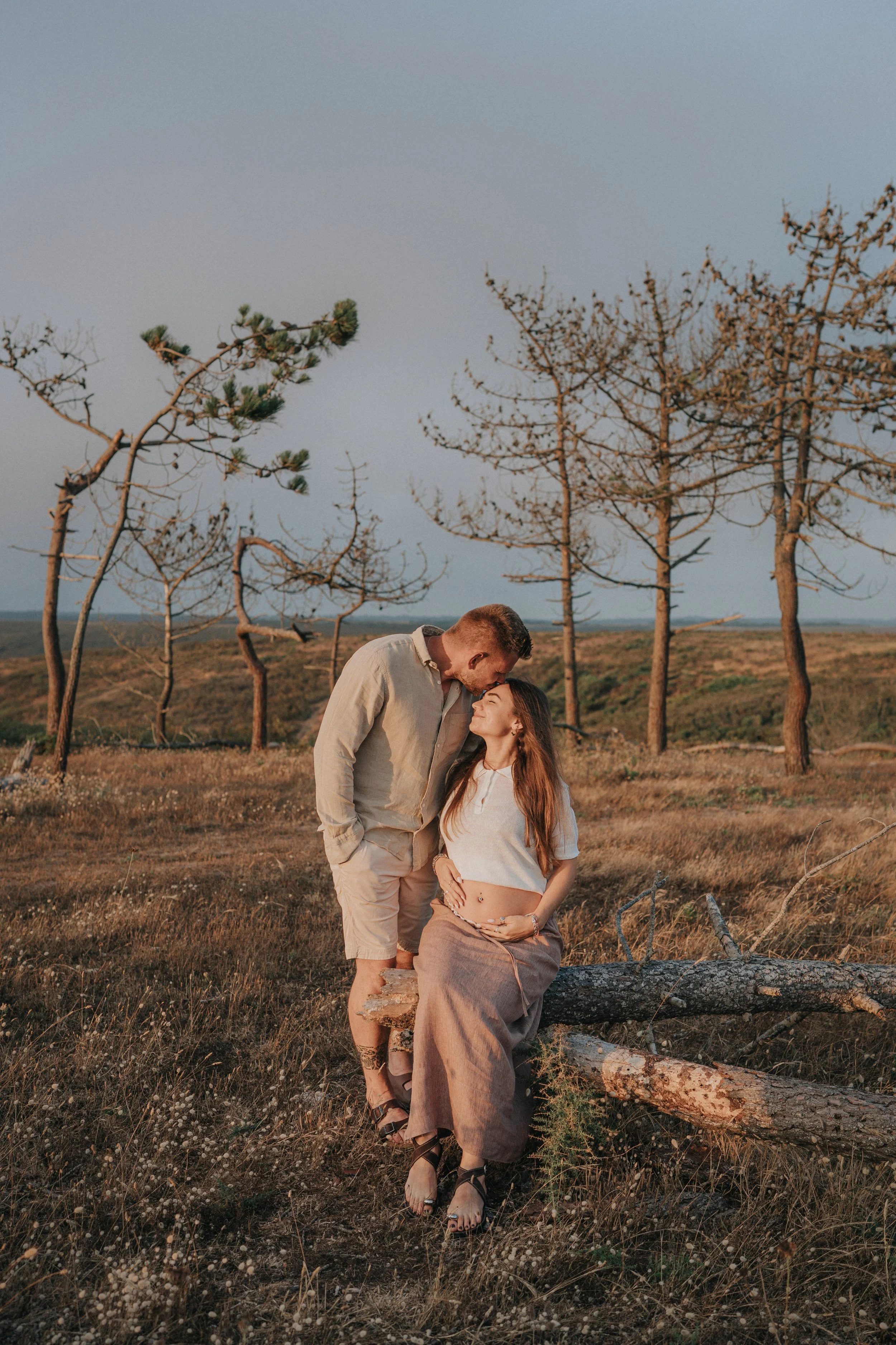 A young man and woman standing in a field of grass and trees, with the man leaning down to kiss the woman on her forehead. The woman is sitting on a log, and both are dressed in light, casual clothing. The scene is bathed in warm, natural light.