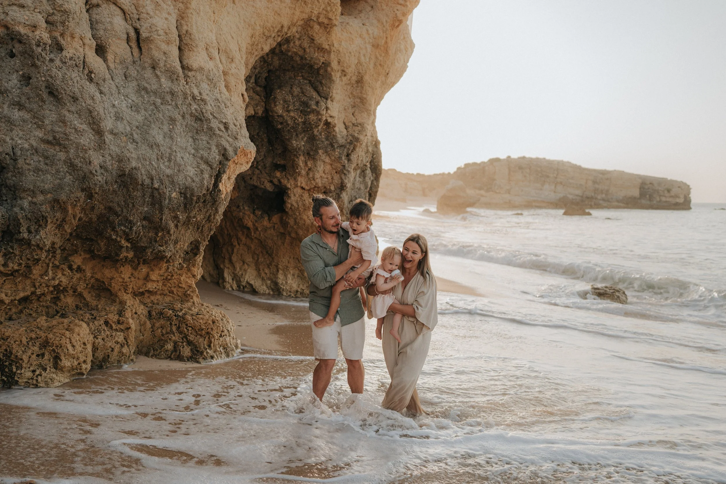 A family of five enjoying a walk by the ocean near rocky cliffs at sunset. The father holds one child, while the mother holds another. The family is smiling and dressed in light clothing, walking in shallow water.