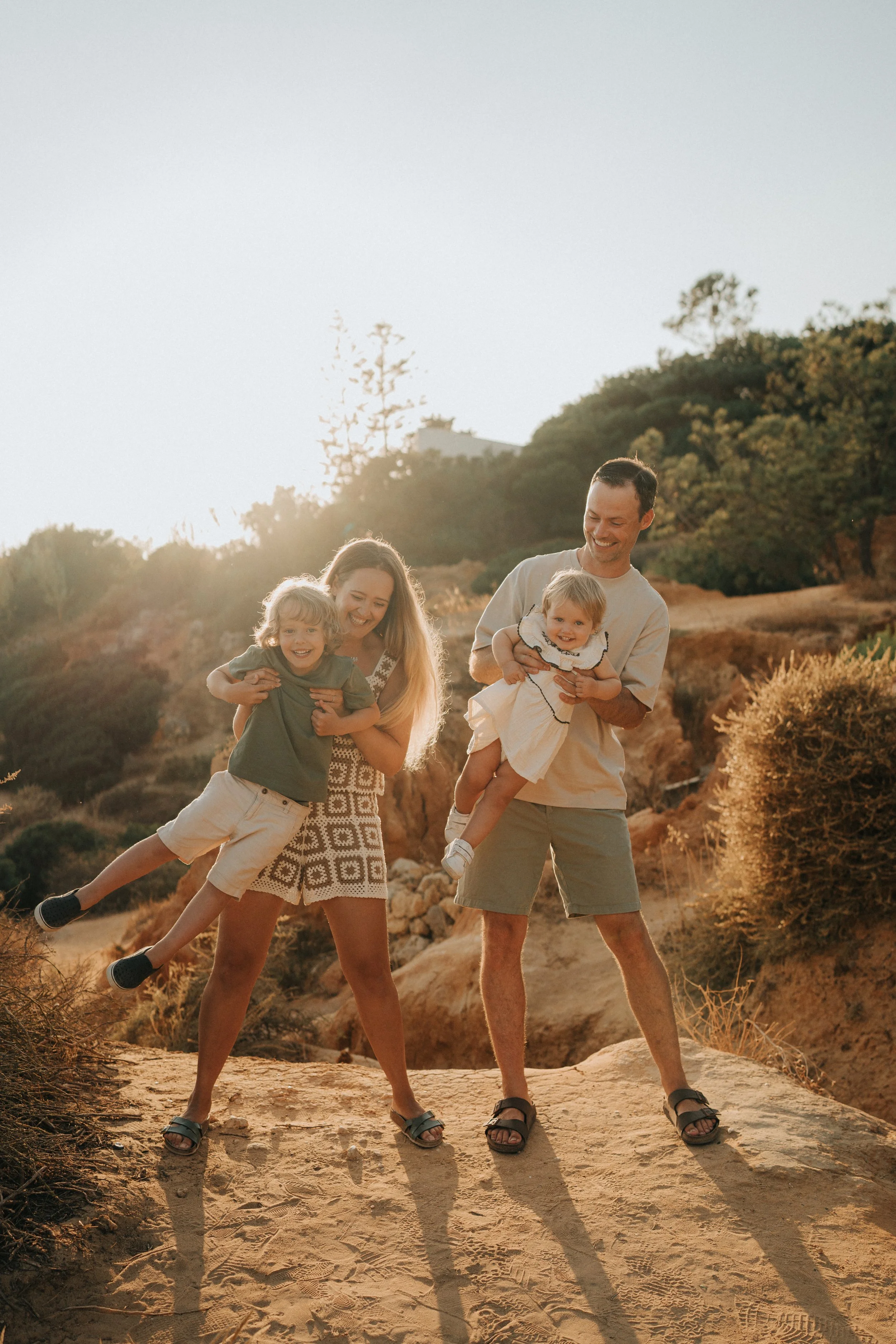 A family of four, two adults and two children, smiling and playing outdoors in a desert landscape during sunset, with trees and rocky hills in the background.