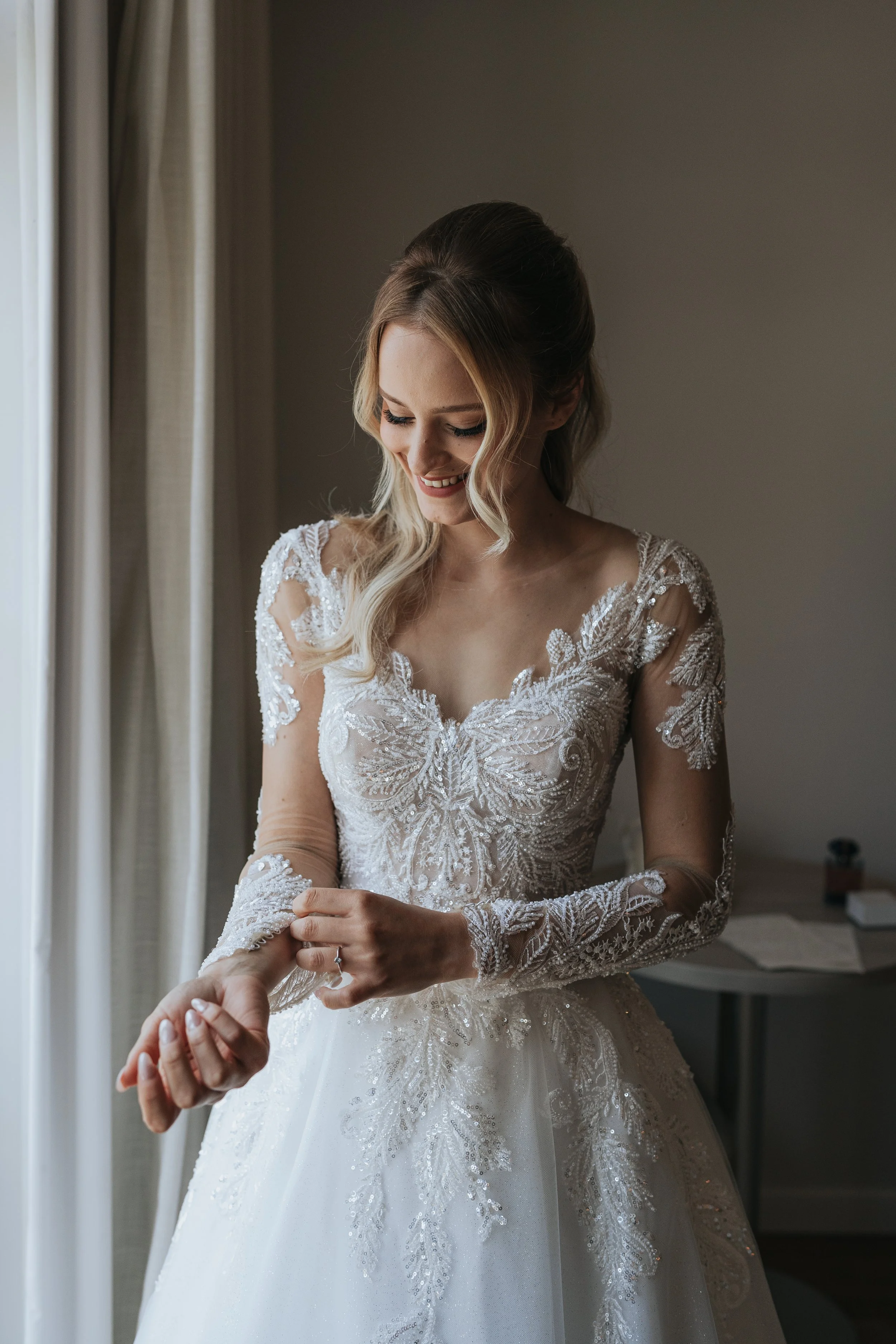 A bride with long wavy blonde hair in a wedding dress with intricate lace and beadwork, smiling as someone puts a ring on her finger, standing near a window with beige curtains.