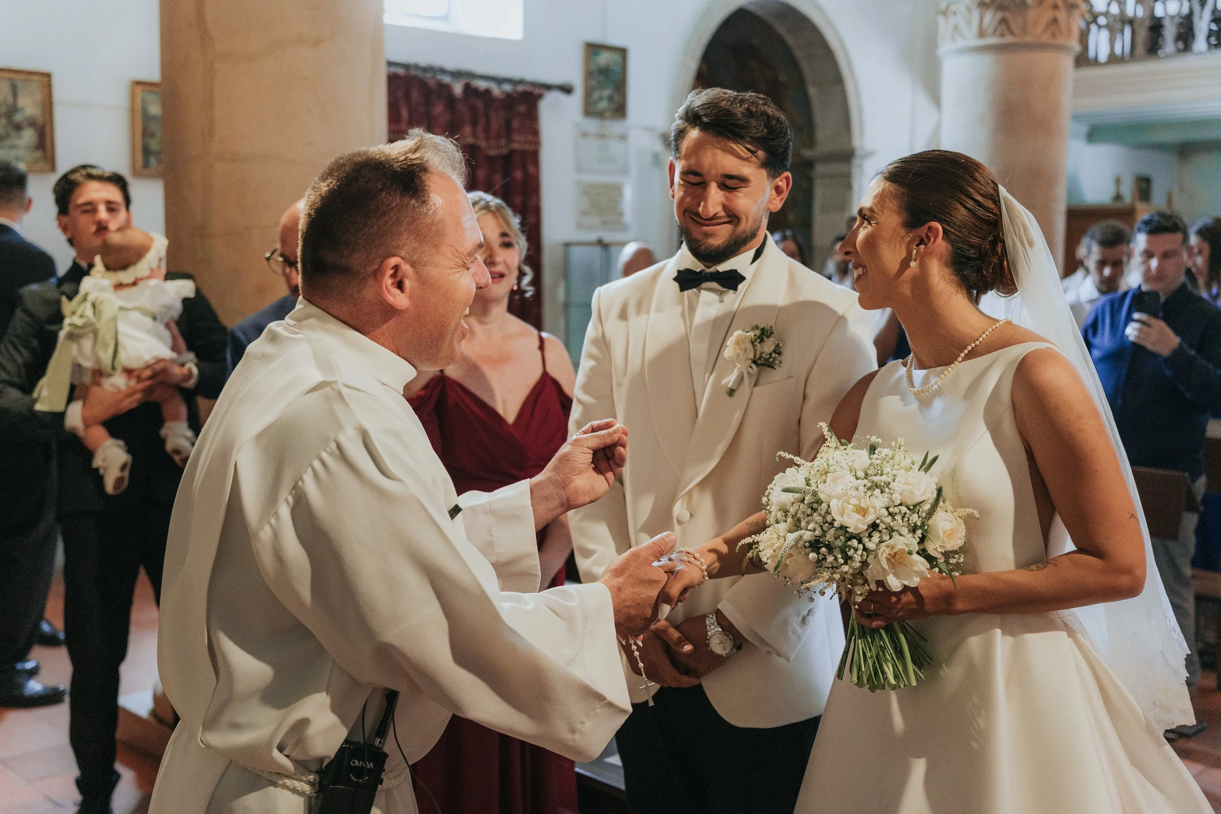 A wedding ceremony with a bride in a white gown holding a bouquet, a groom in a tuxedo, and an officiant exchanging vows inside a church with guests in the background.