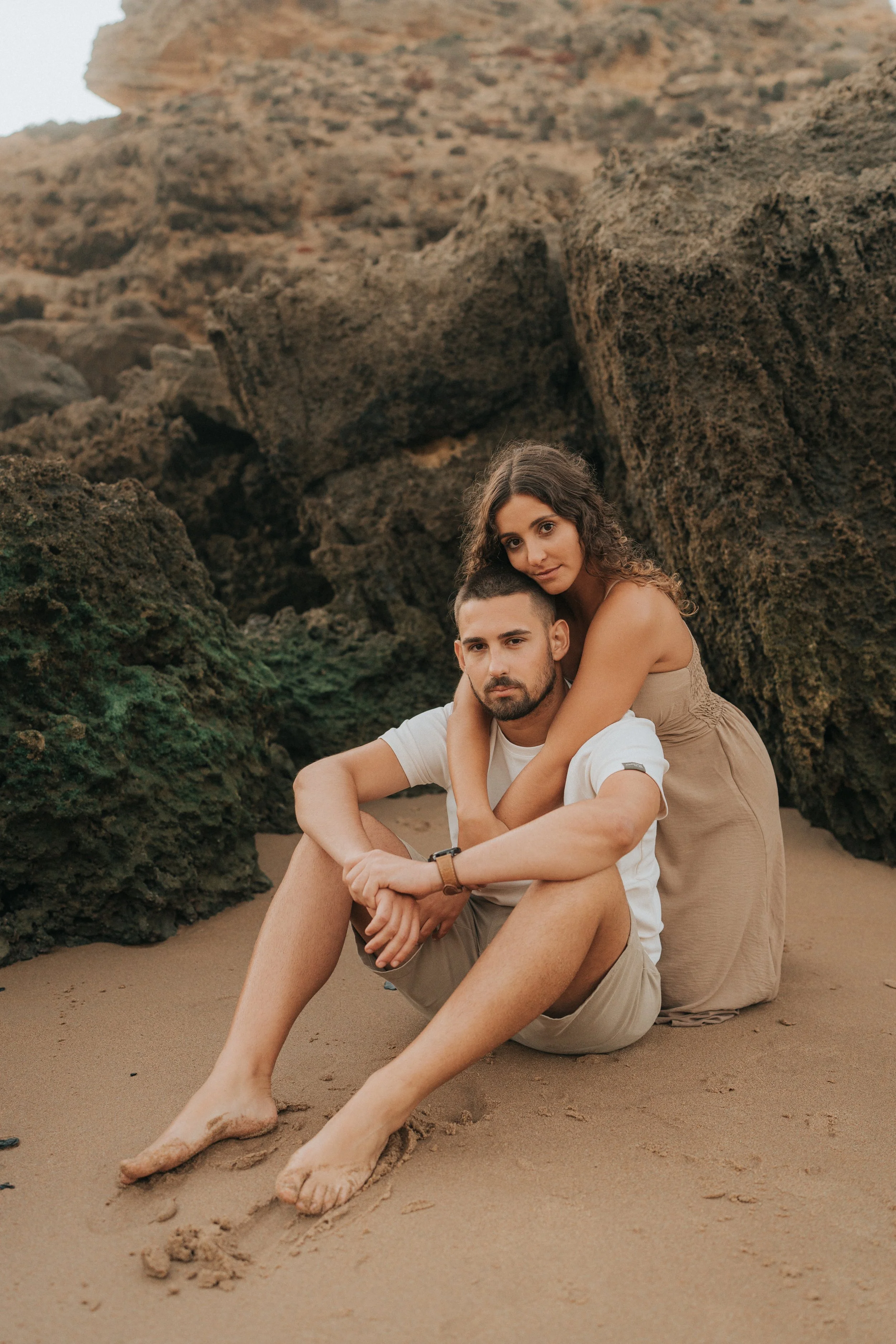 A young couple sitting on the beach, with the woman hugging the man from behind, surrounded by large rocks and sandy terrain.