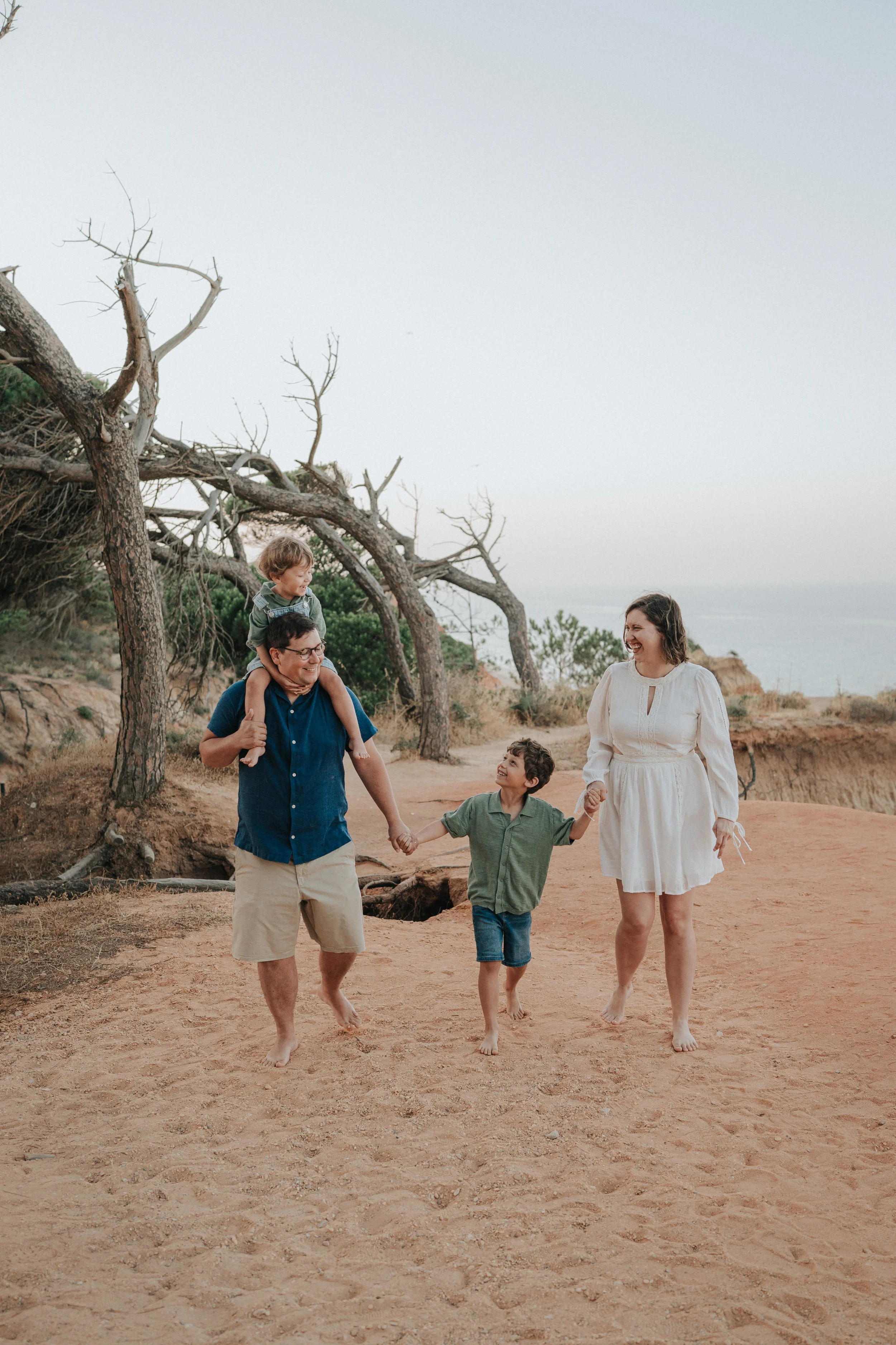 A family of five walking along a sandy outdoor trail near a coastal area with dry trees, with a cloudy sky.
