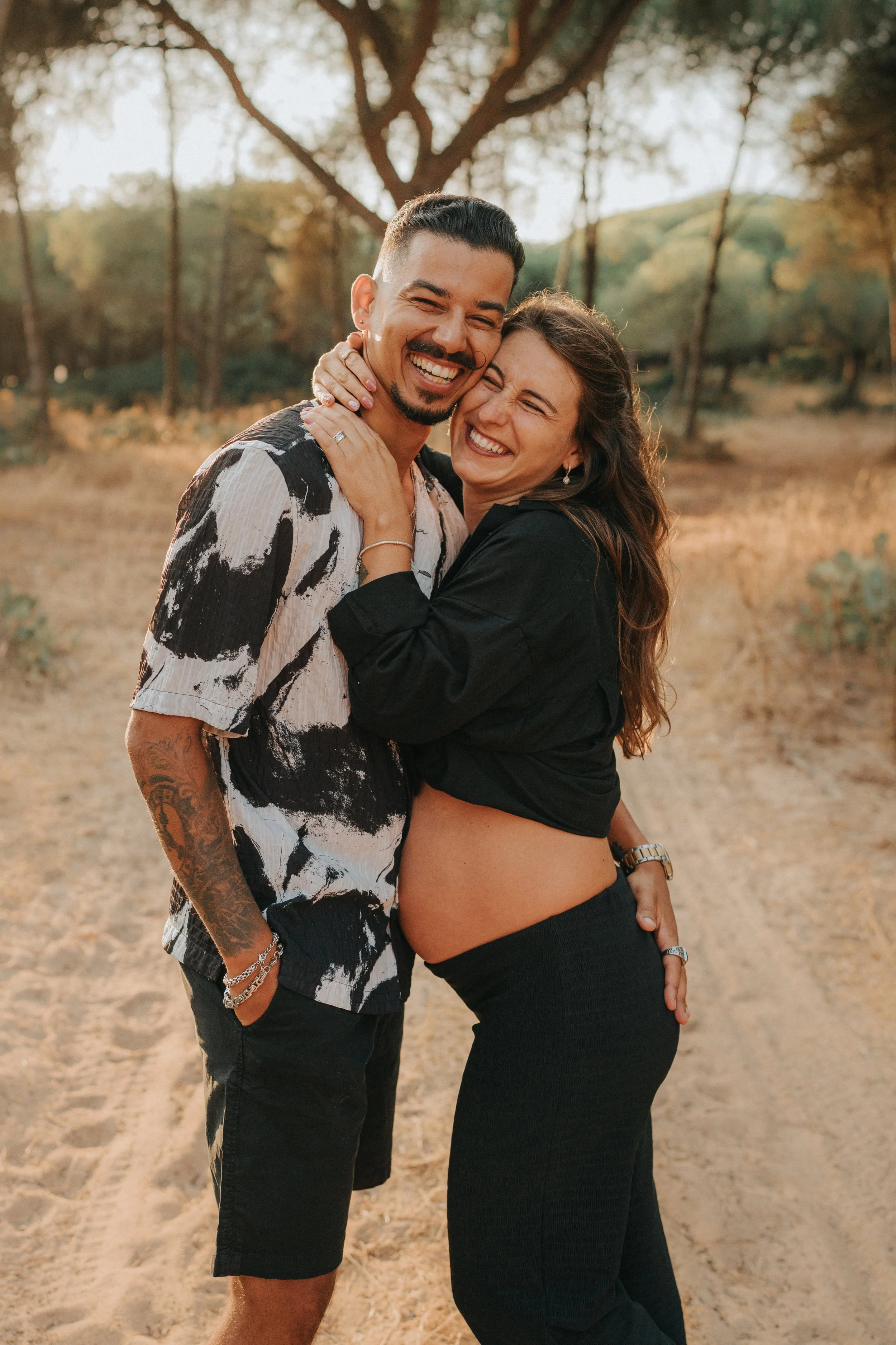 A man and woman are embracing and smiling outdoors on a dirt trail with trees in the background, during sunset.