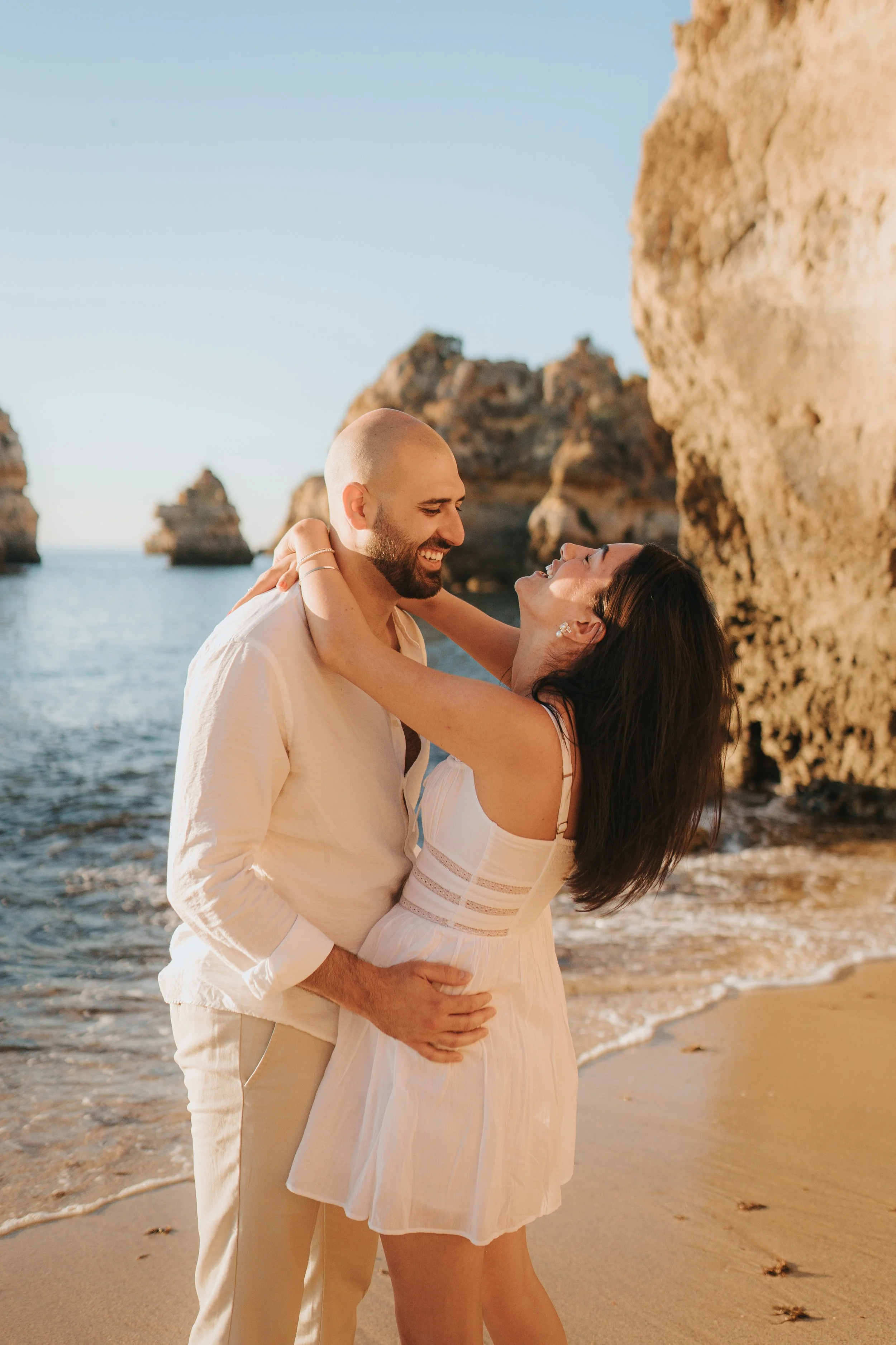 A smiling couple dancing by the shoreline with rocky cliffs in the background, during sunset.