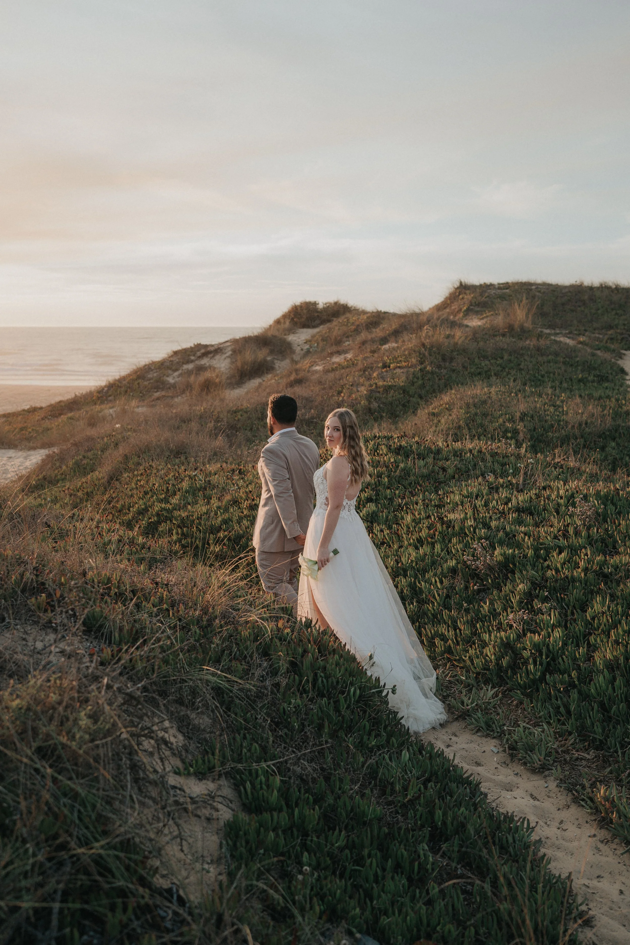 A bride and groom walking on a sandy beach with dunes and vegetation in the background during sunset.