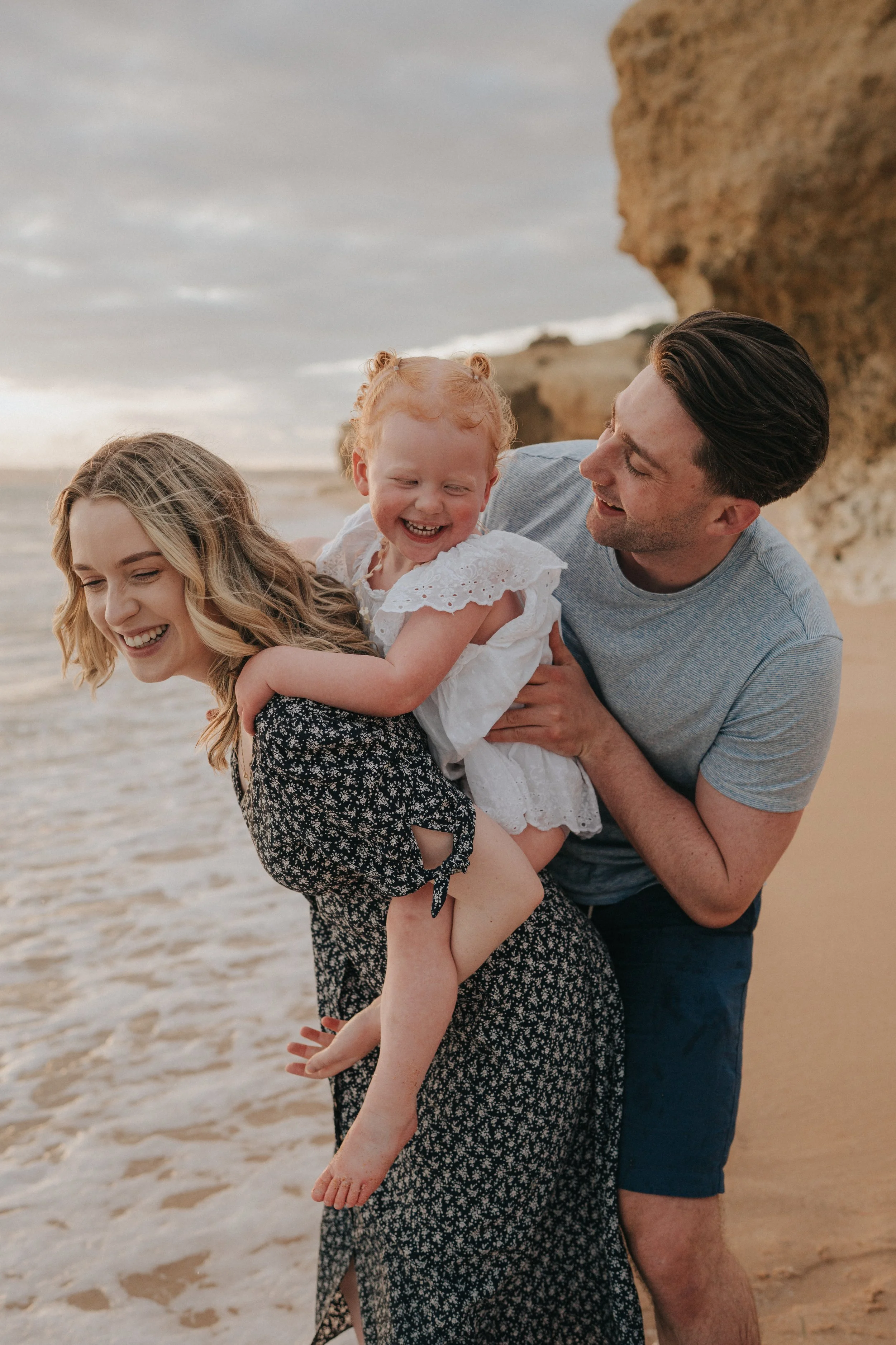 A happy family on the beach, with a woman holding a young girl, while a man supports the girl from behind. They are all smiling and laughing, enjoying a moment together near the ocean with cliffs in the background.