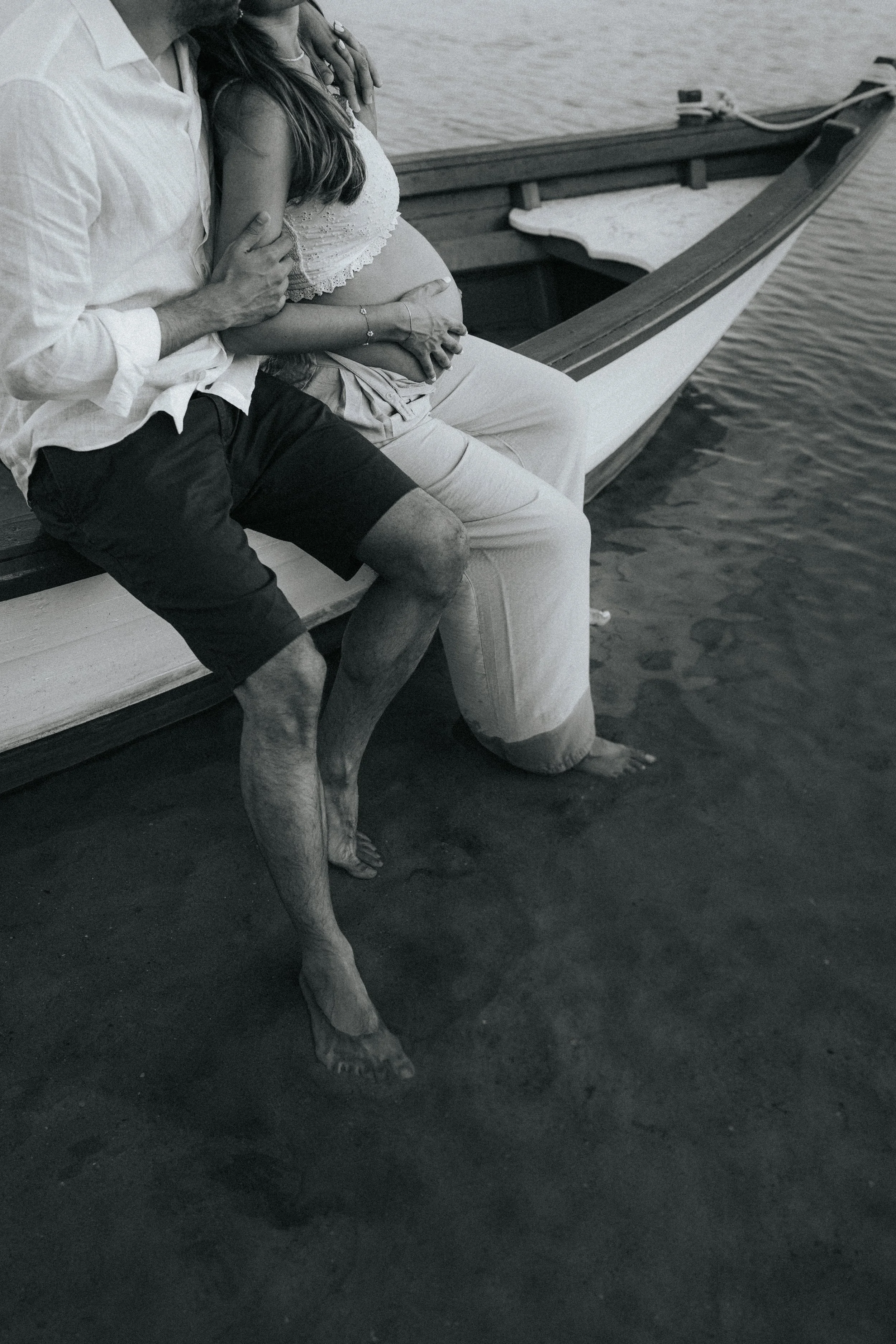 A person with a shirt and shorts sitting beside another person with a pregnant belly, all near a boat at the water's edge in black and white.