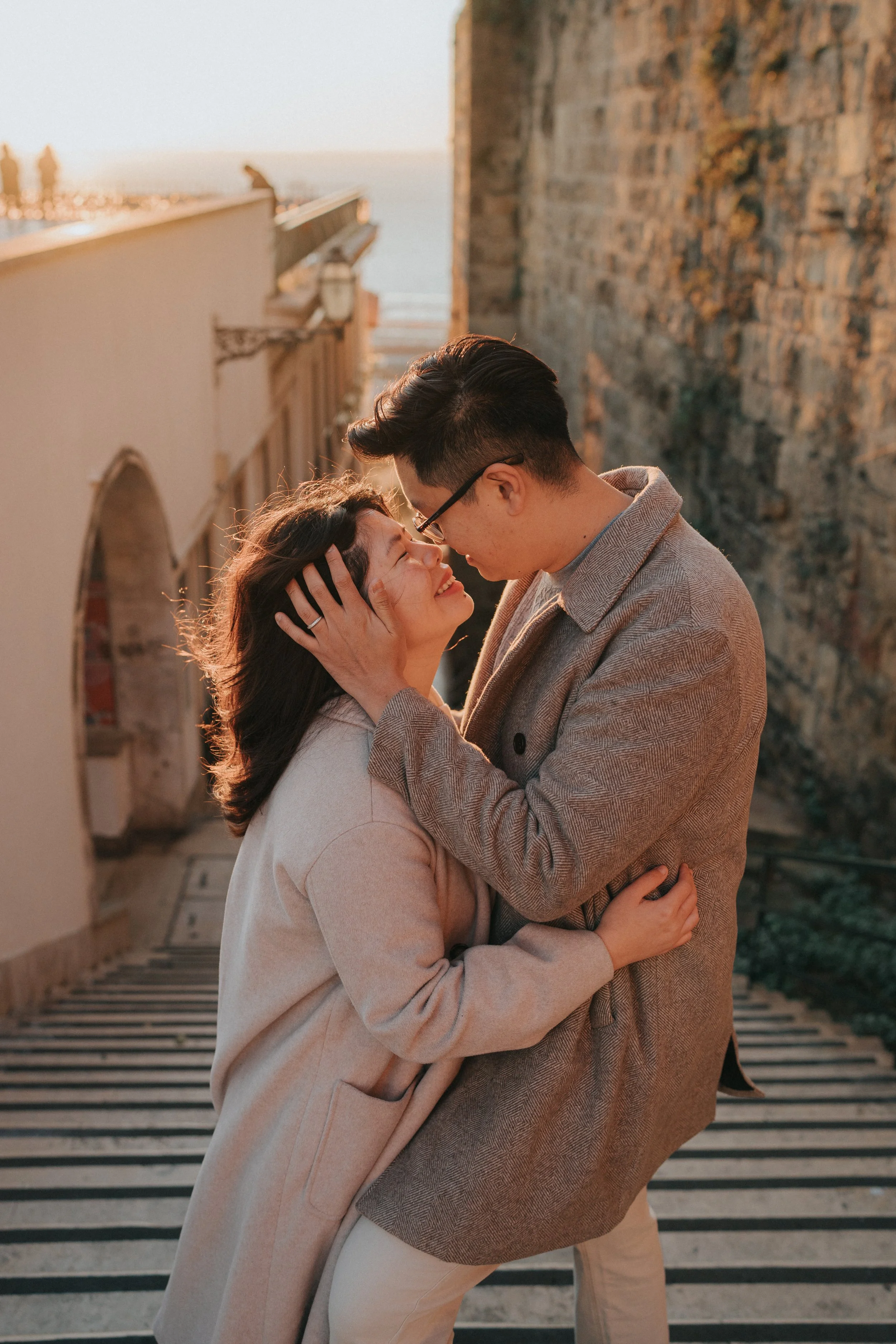 A man and woman are embracing each other outdoors during sunset on a staircase near a stone wall, smiling warmly at each other.