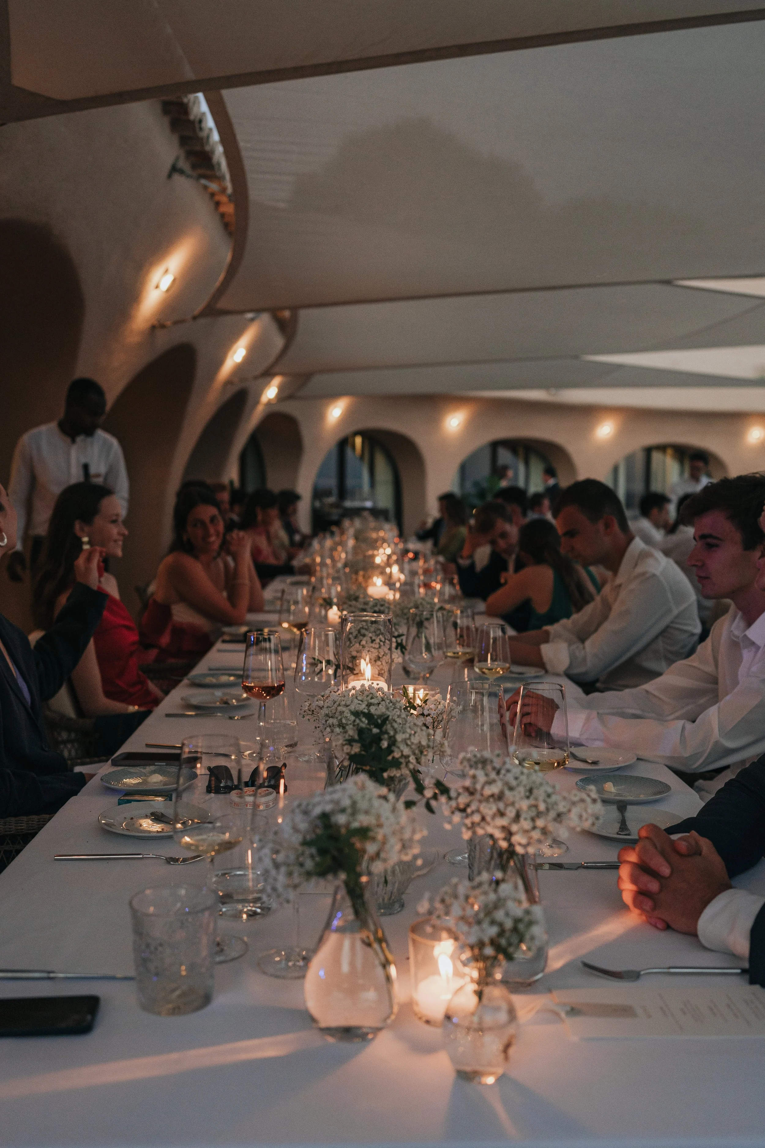 A long formal dinner table set with white tablecloths, flowers, candles, and glassware, with guests seated on both sides, in a dimly lit room with arched windows and warm lighting.