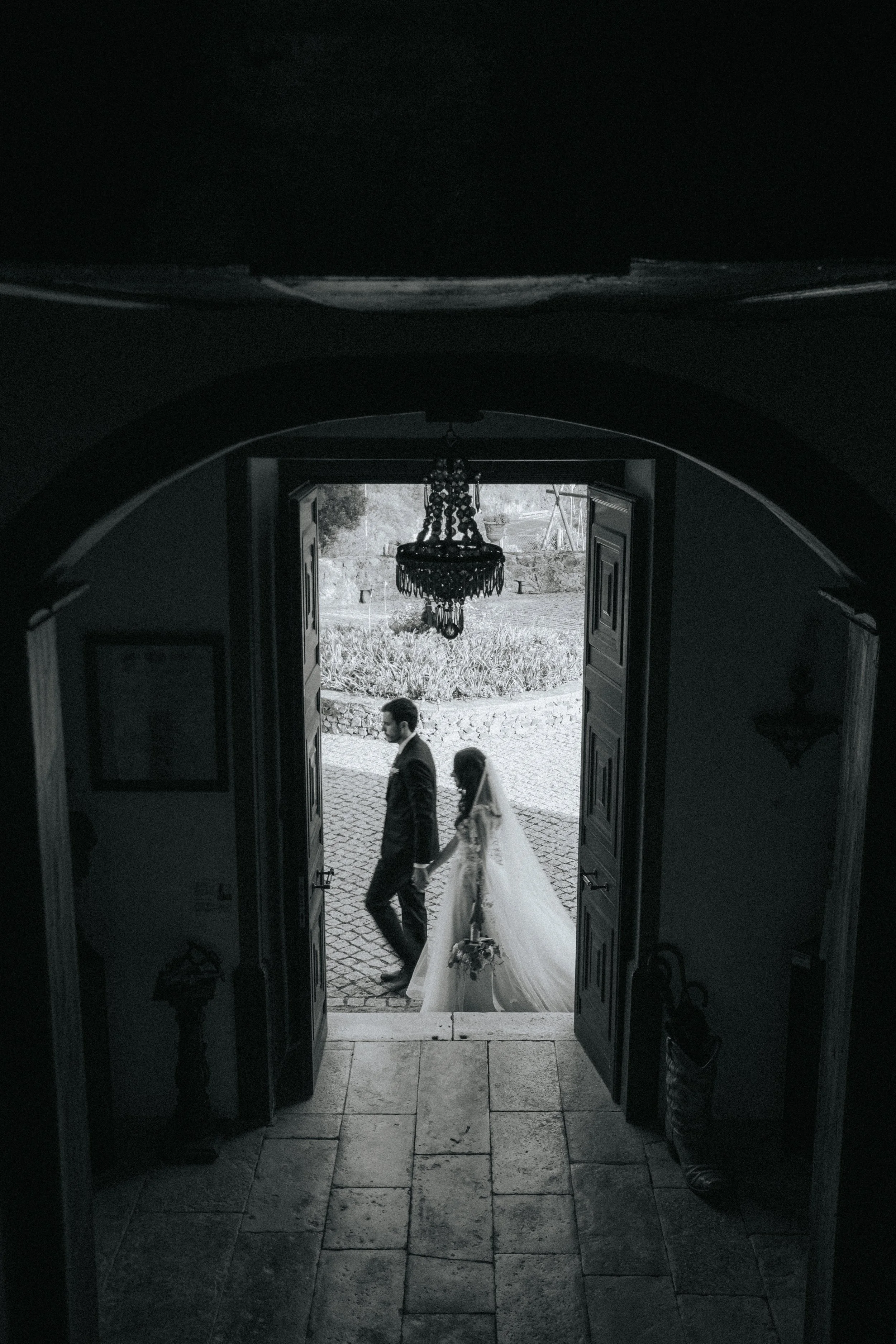 A man and woman walking outside through a doorway, with the woman wearing a wedding dress, indicating a wedding or formal event.